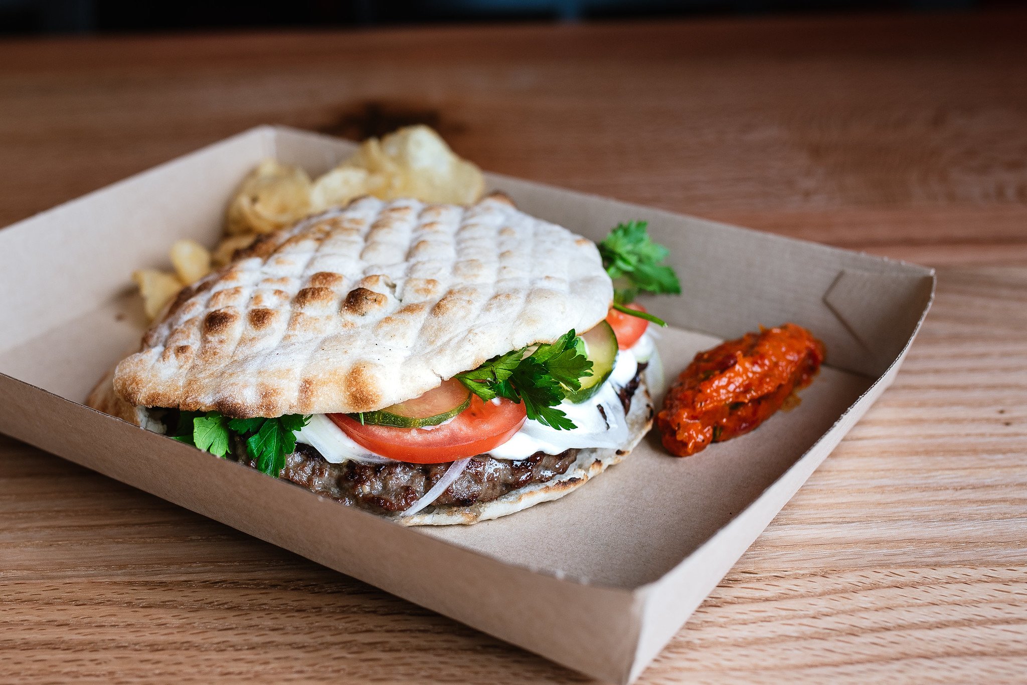 A grilled pita sandwich with beef, sliced tomato, cucumber, onion, parsley, and sauce, served with potato chips and a side of red spicy sauce in a cardboard tray on a wooden surface.