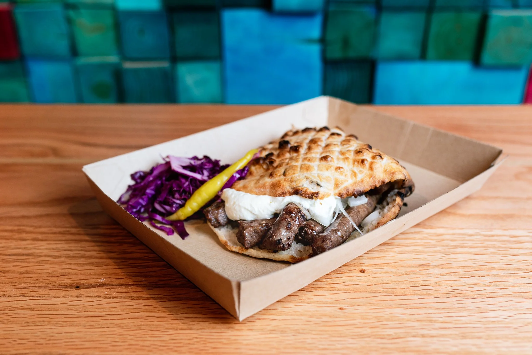 Sandwich with grilled meat, cheese, and a toasted flatbread, served with purple cabbage and a yellow chili pepper on a cardboard tray.