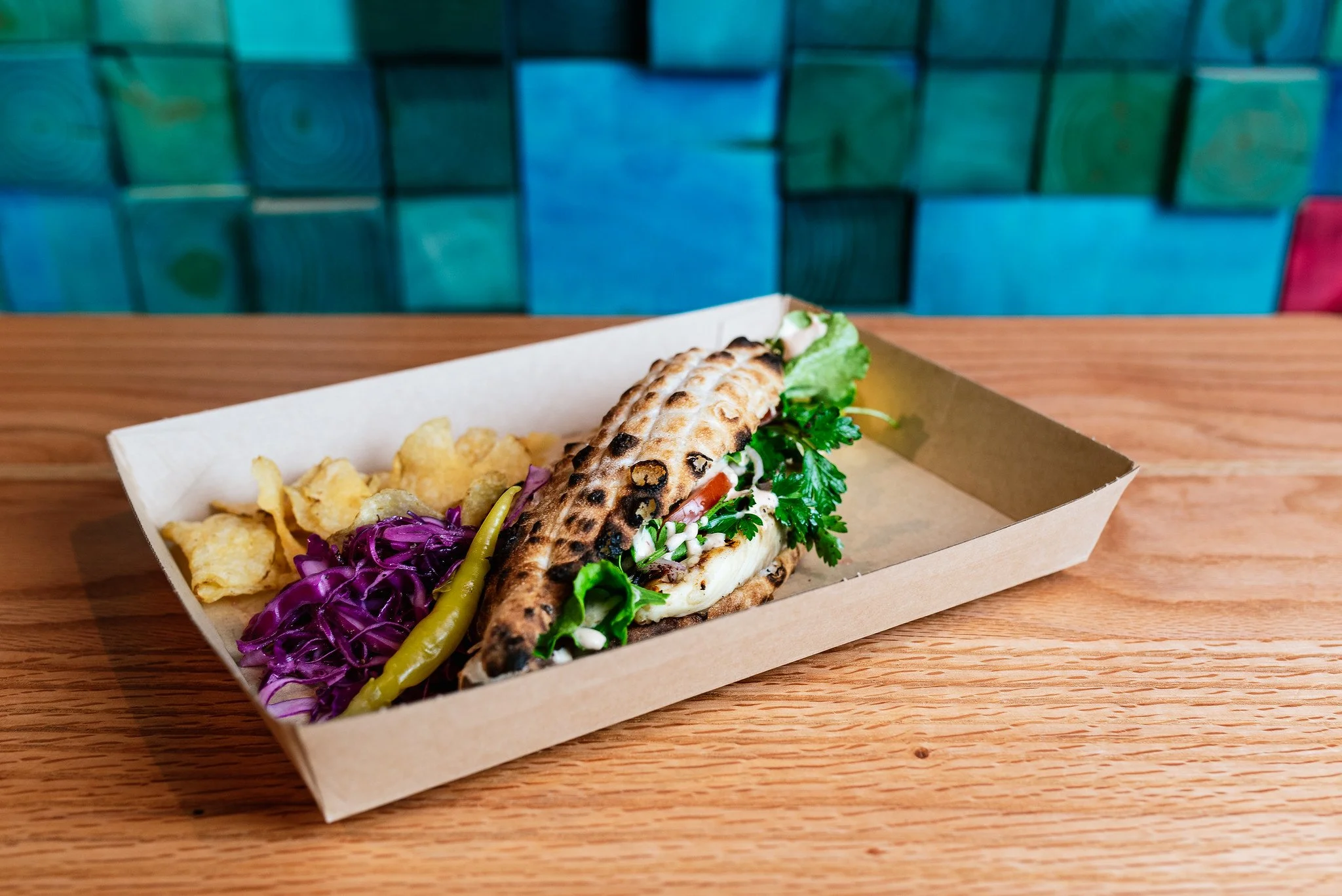 A food tray containing an Asian-style grilled sandwich, purple cabbage, potato chips, and a green chili pepper on a wooden table.