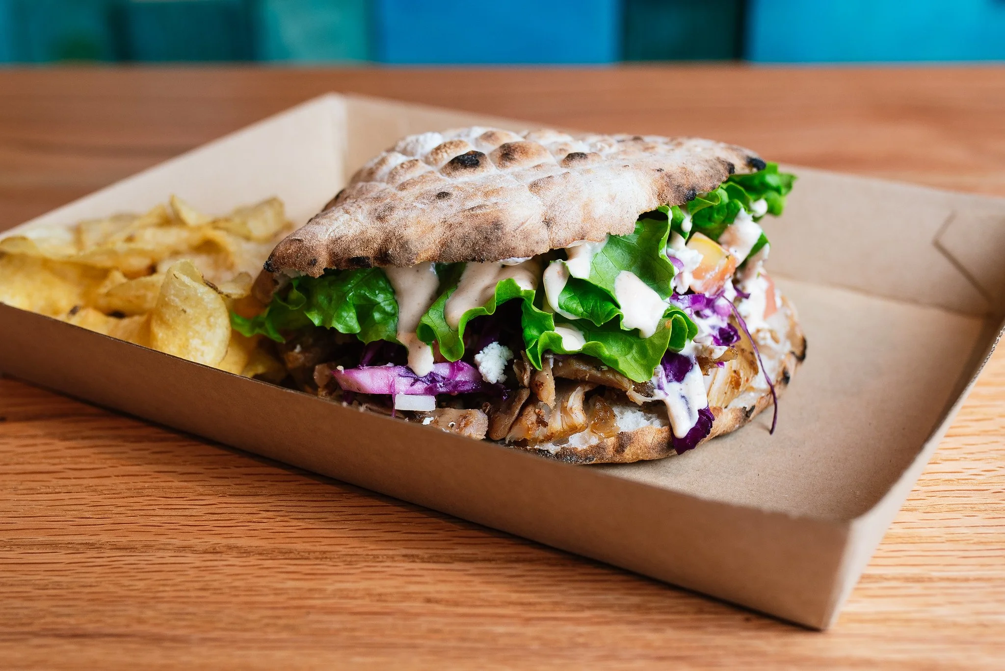 Giant sandwich with grilled flatbread containing lettuce, purple cabbage, shredded meat, and creamy dressing, served with potato chips in a cardboard tray on a wooden table.