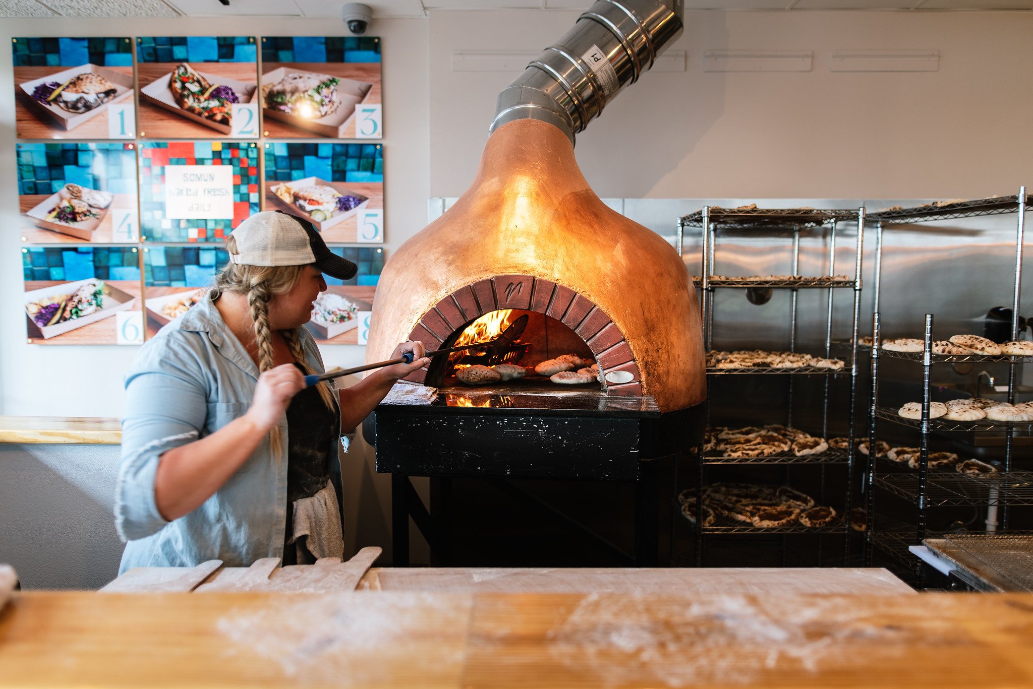A woman wearing a baseball cap and apron baking cookies in a brick oven, with shelves of cookies on racks beside her and a colorful display of menu images on the wall behind.