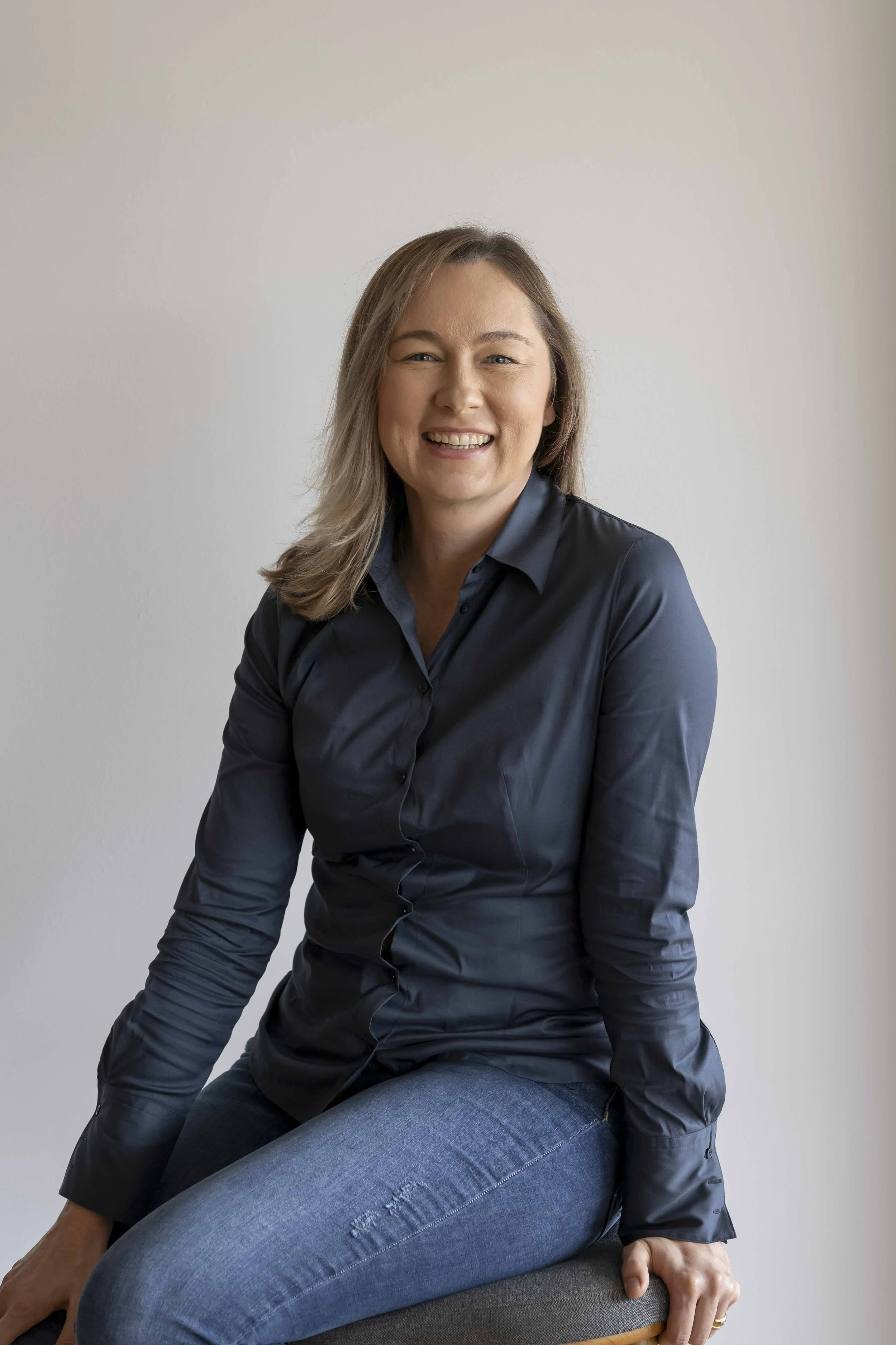 A woman with shoulder-length blonde hair, wearing a dark blue satin blouse and blue jeans, smiling and sitting on a stool against a plain light-colored wall.