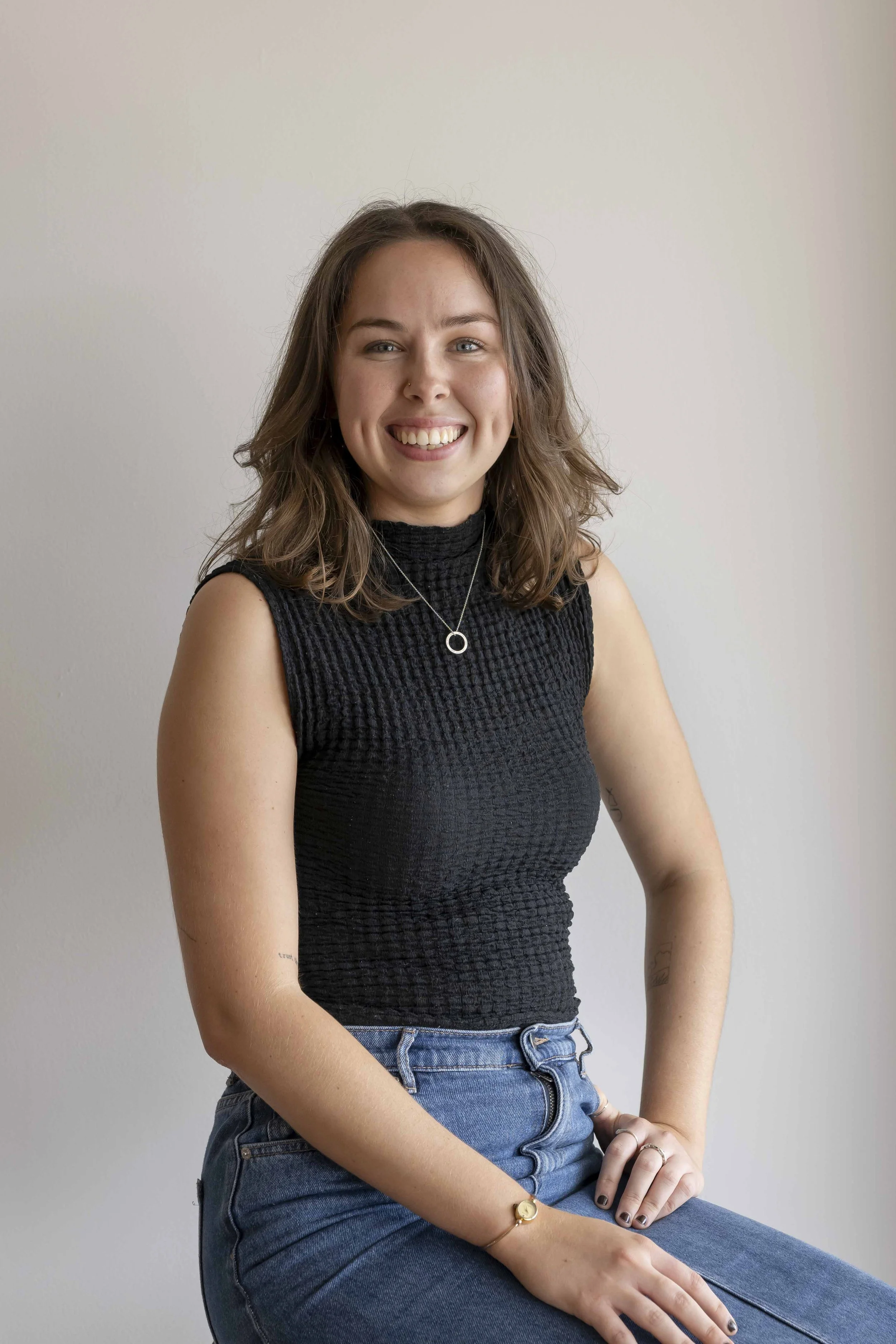 A young woman with shoulder-length brown hair, wearing a black sleeveless textured top and blue jeans, sitting against a plain light-colored wall, smiling at the camera.