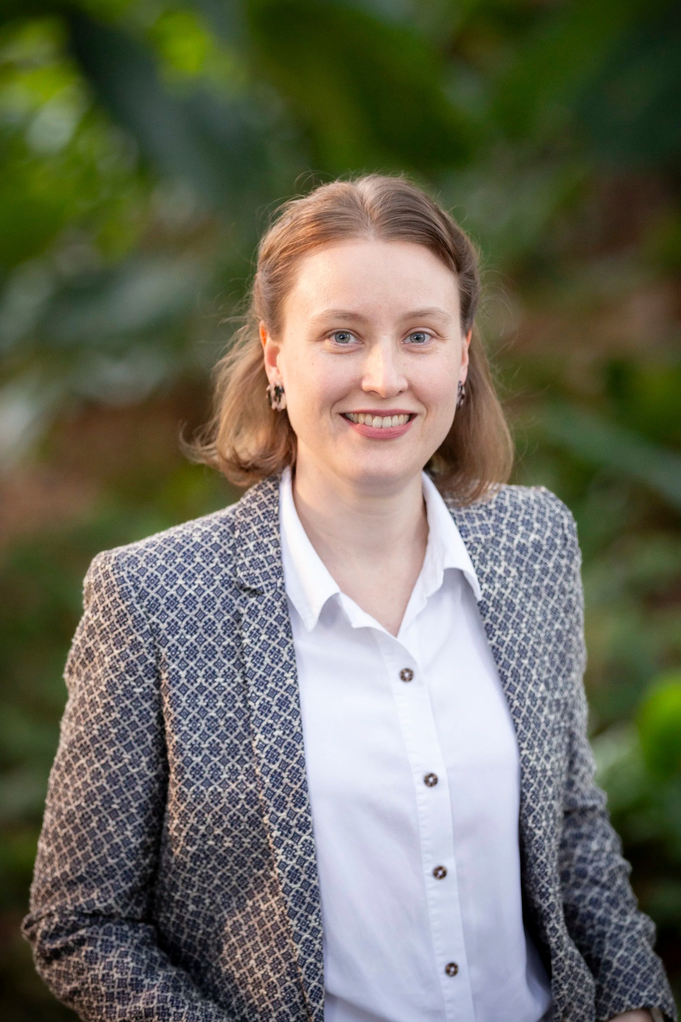 Portrait of a woman with short brown hair, wearing a white blouse and patterned blazer, smiling outdoors with blurred greenery in the background.