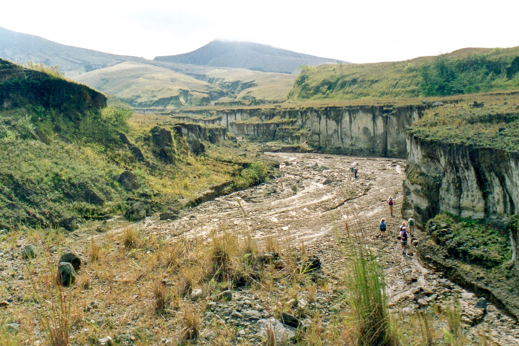   Sur ce cliché, où notre file indienne donne l’échelle du ravin, on trouve les 3 éléments caractéristiques de la topographie des flancs externes du volcan : le ravin entaillé dans le matériel remanié en terrasse ; la bosse d’une coulée pyroclastique issue de l’éruption (à droite. ; les interfluves matelassés par les cendres et entaillés de ravins (à gauche). Tout à l’arrière-plan, un des 2 cratères annexes anciens et son dôme dans le nuage.  