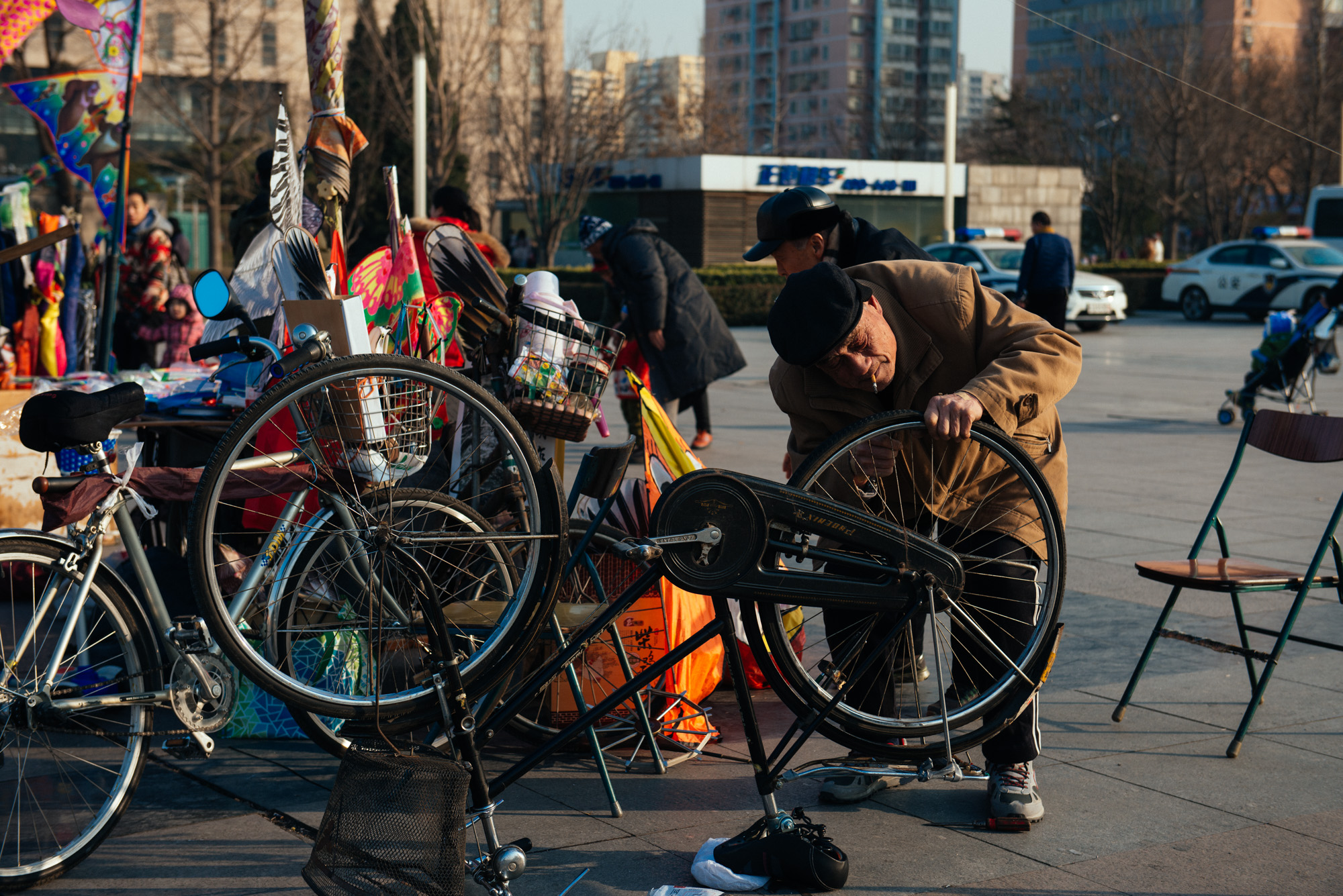 Beside the guy that made and sold kites, an old man fixes the wheel of his bicycle.