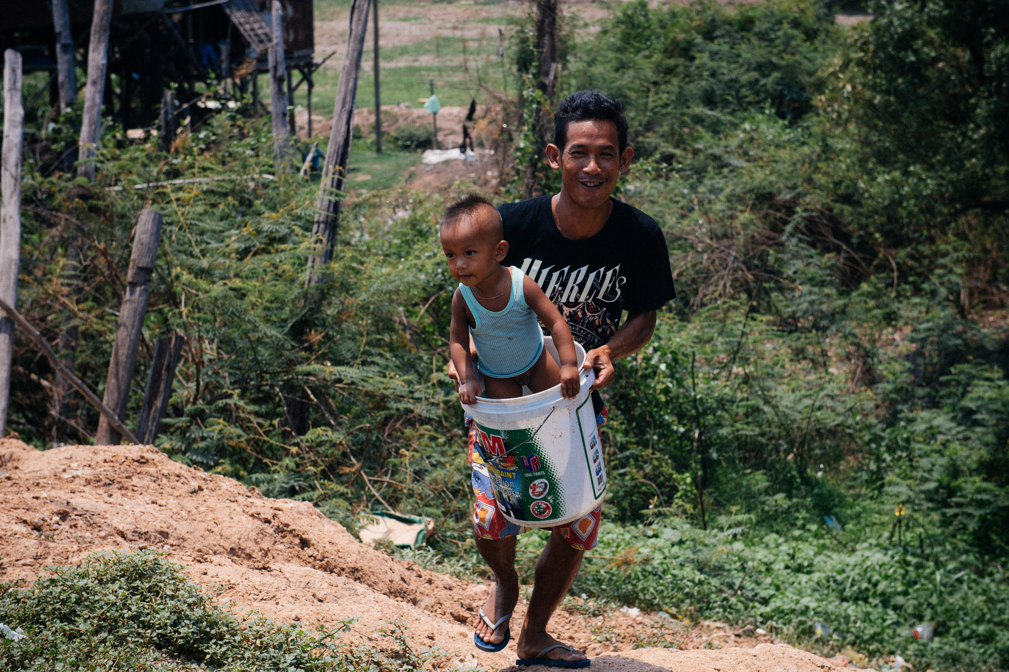 Coming up the hill, from the waterbed, a man carries a boy inside a bucket. The boy loves the game.