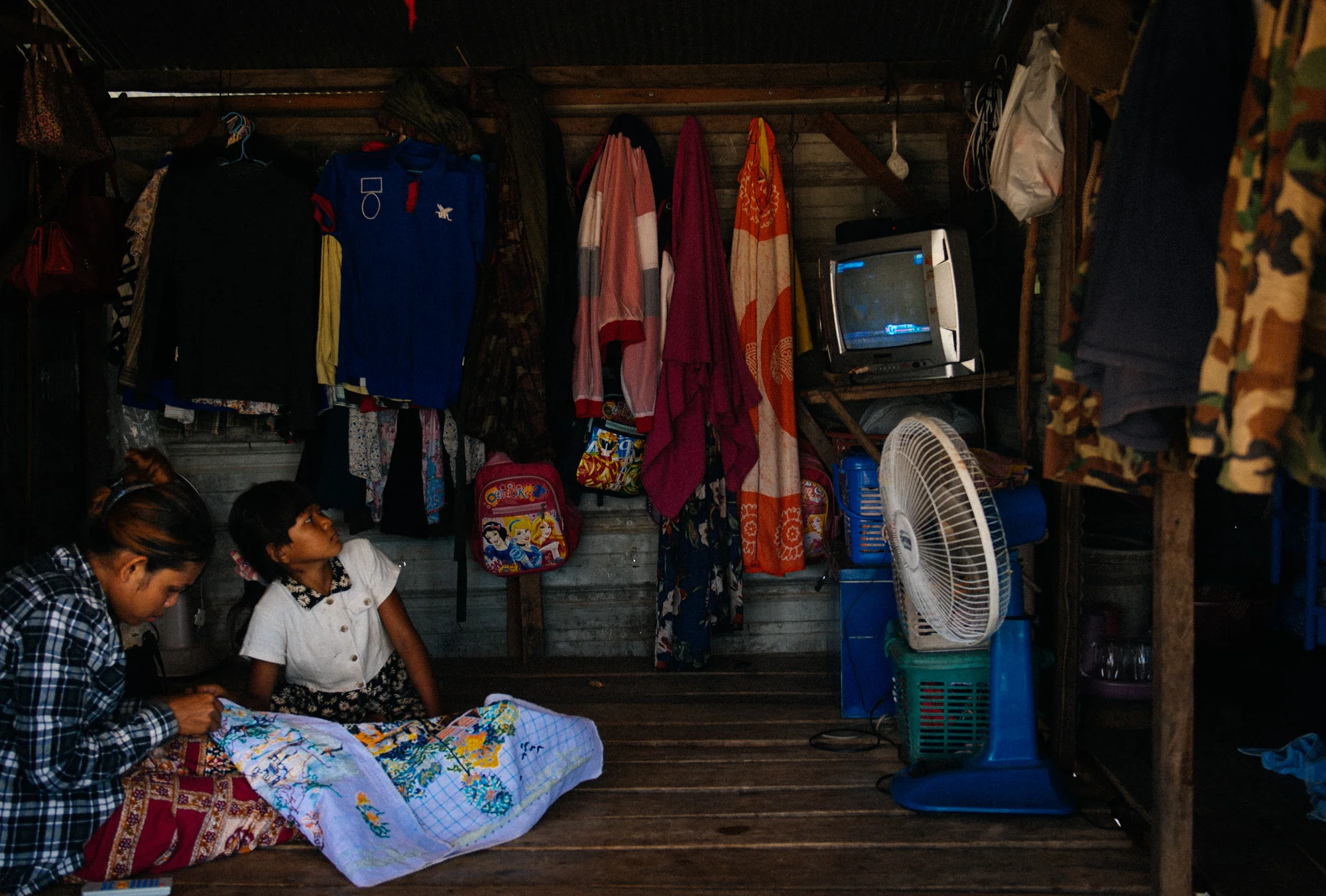 Girl watches television while another sews inside their house. Like this place, most shacks have only one room where several people sleep either on the ground or in hammocks.
