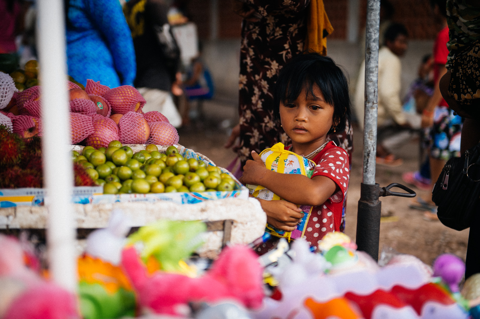 Little girl with something in her eye clings to her backpack as she browses fruit at a market across the street from the children's hospital.