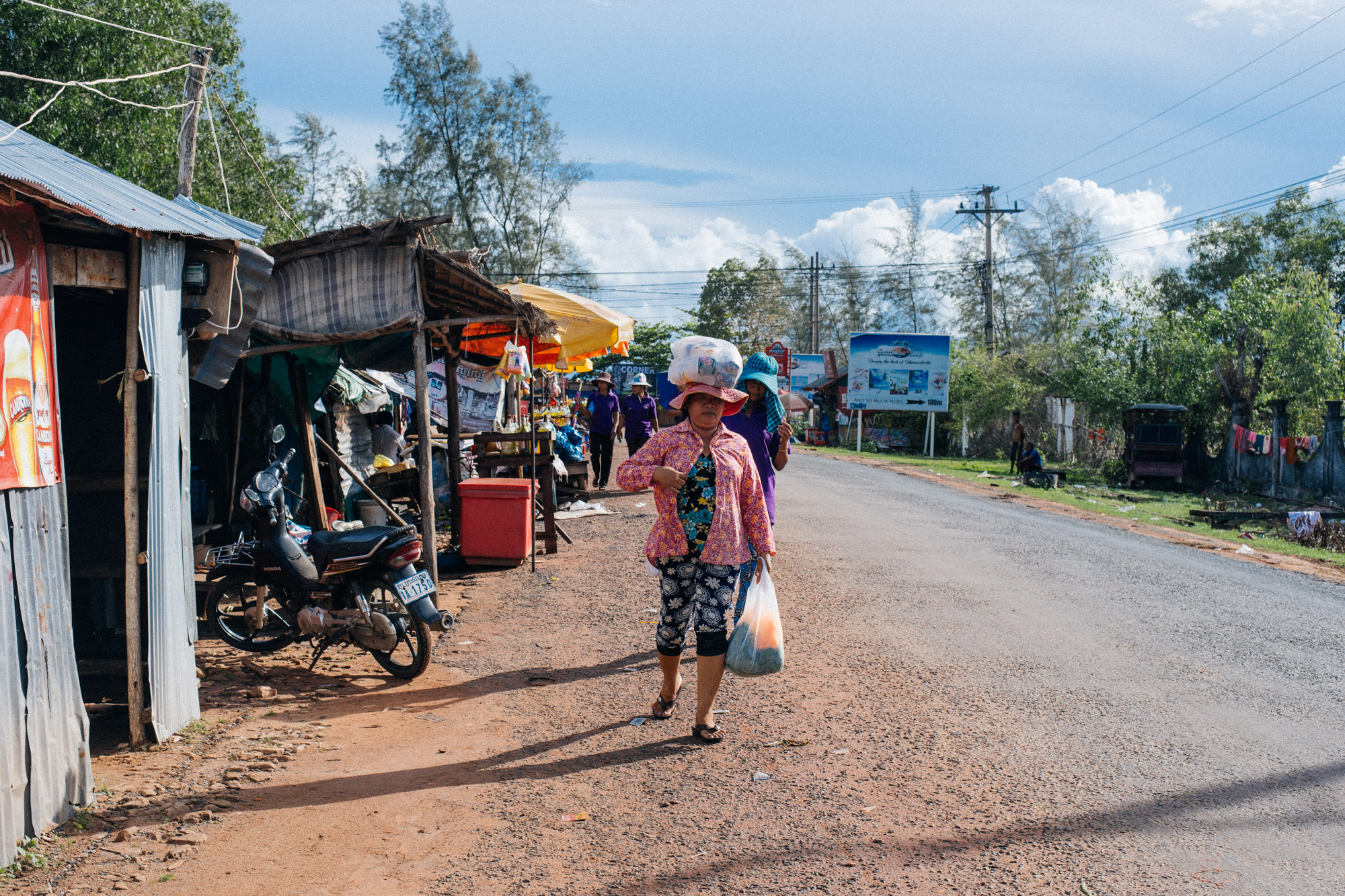 Woman walks past carrying groceries balanced on her head.