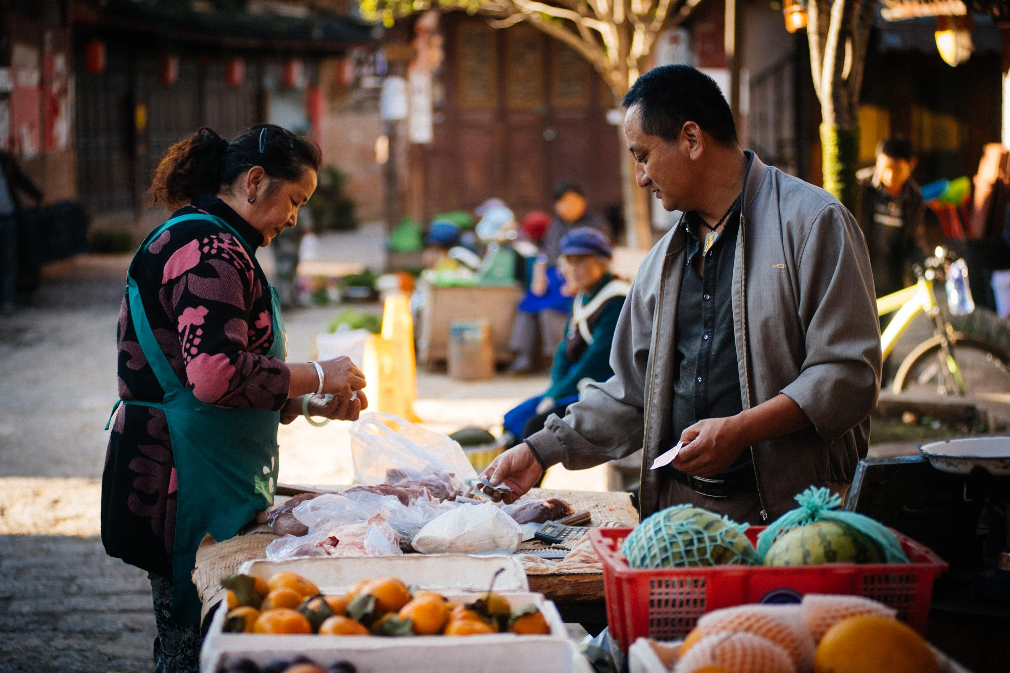 Butcher sells a chunk of pork to the woman that is selling vegetables in the stand right beside him. I can only assume she got the best part as she was the day's first customer.