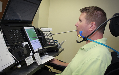 Denny, sitting at an adjustable height desk, uses a mouth stick and an iPad to operate a laptop at Guardian Fence Suppliers. Denny is known in the office as the “Zoho Czar,” referring to the Zoho customer relationship management software which he uses and trains other staff on how to use.