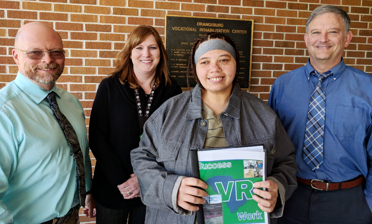 Melissa Freidman, former client, displays the book she created documenting her experiences at VR. Around her are (left to right) John Bates, Job Coach; Nicole Kilgus, Counselor; and Stephen Kesler, Vocational Assessment and Career Specialist.