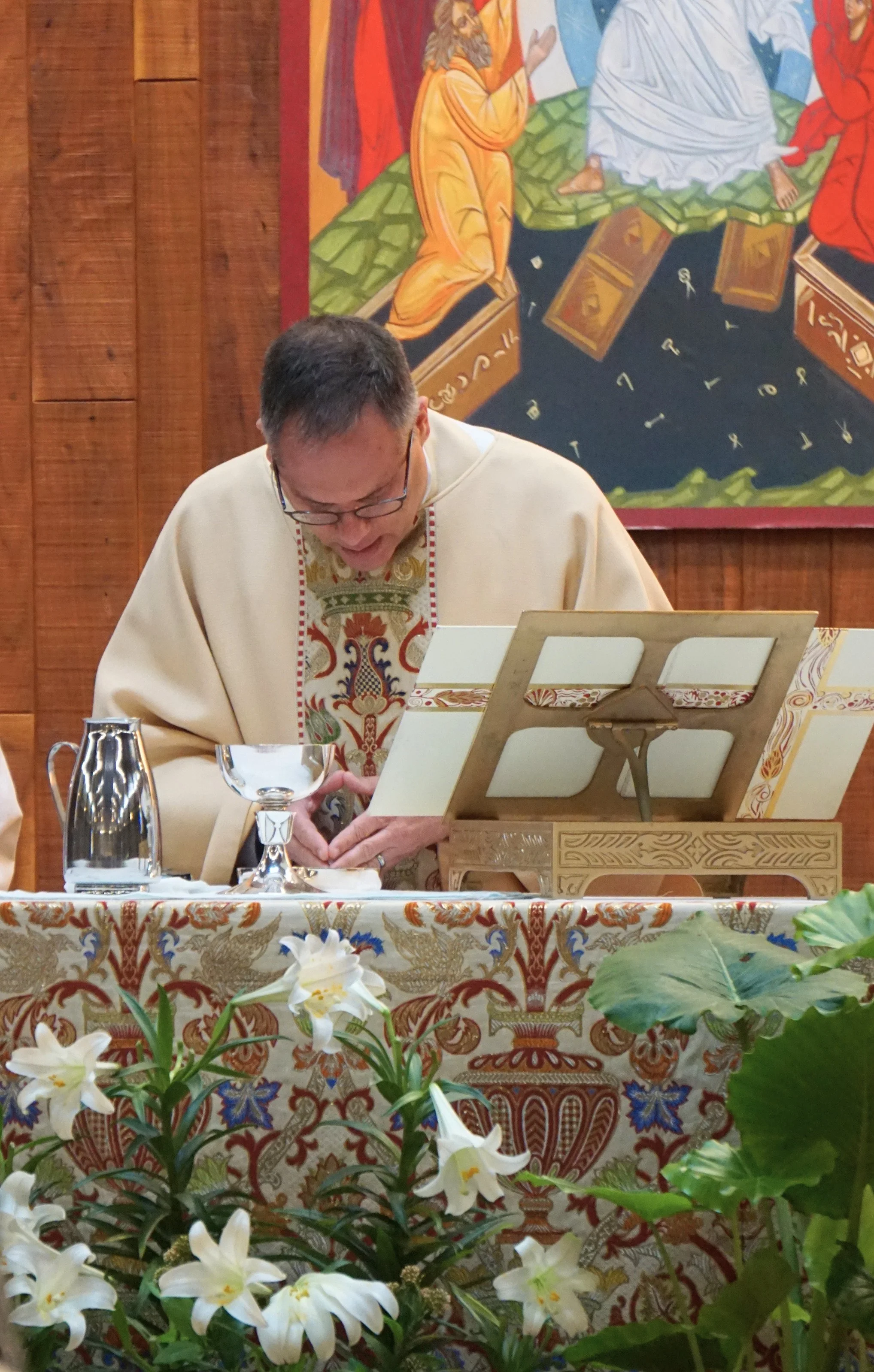 A priest in cream-colored robes with ornate embroidery is standing at an altar decorated with a floral cloth. He is looking down, possibly reading or praying. In front of him is a table with a chalice, a pitcher, and a book stand. The background feat