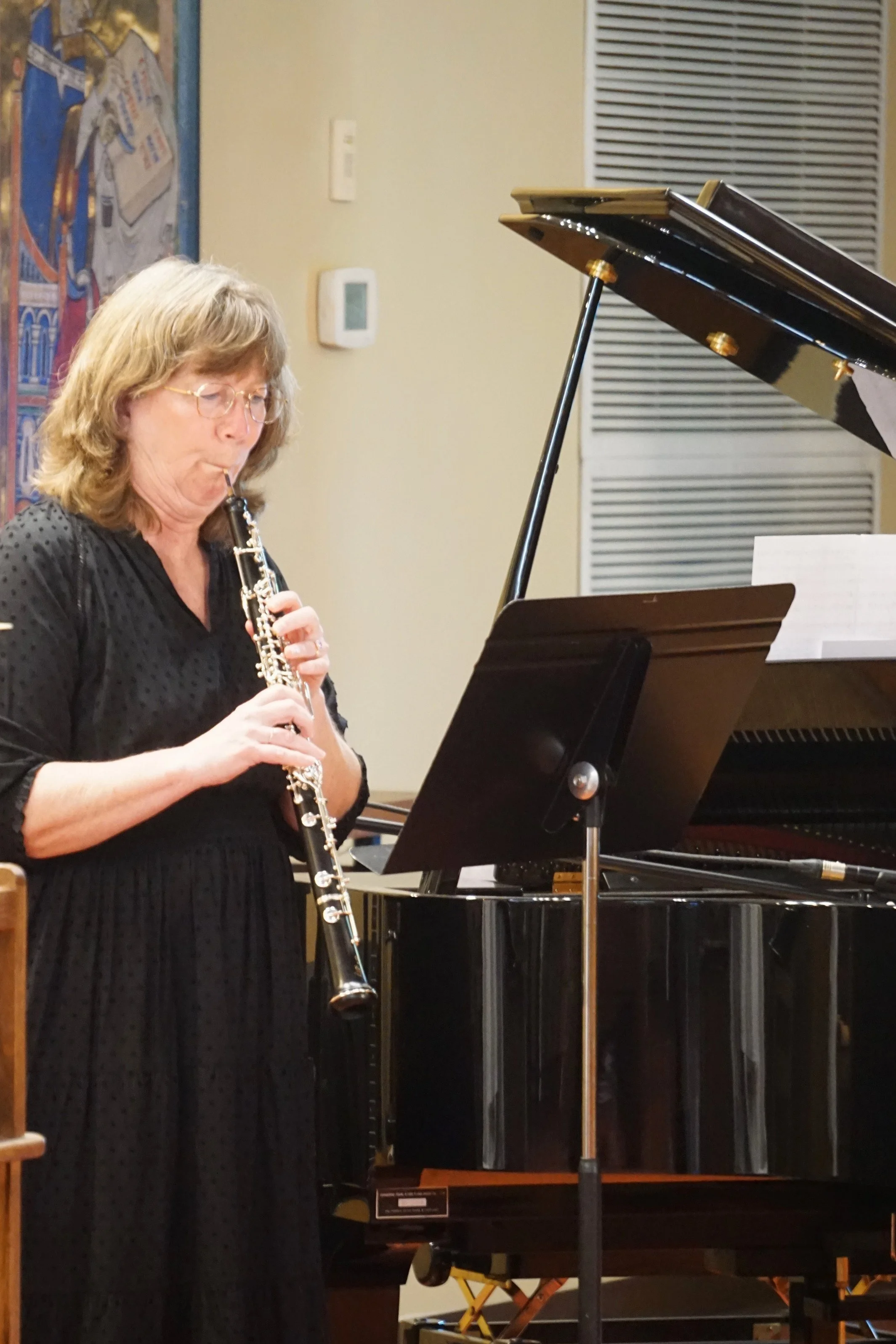 A woman with shoulder-length blonde hair and glasses is playing a clarinet. She is wearing a black dress with a subtle pattern. There is a grand piano and a music stand in front of her. The background includes a wall with artwork, a vent, and thermos