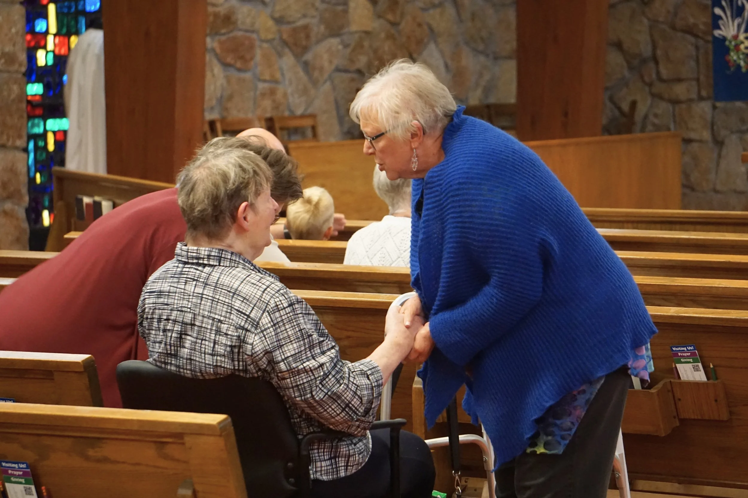 A woman in a blue sweater is kneeling and holding hands with a man seated in a church pew, with other people seated nearby, inside a church with stained glass windows and house-like wood pews.