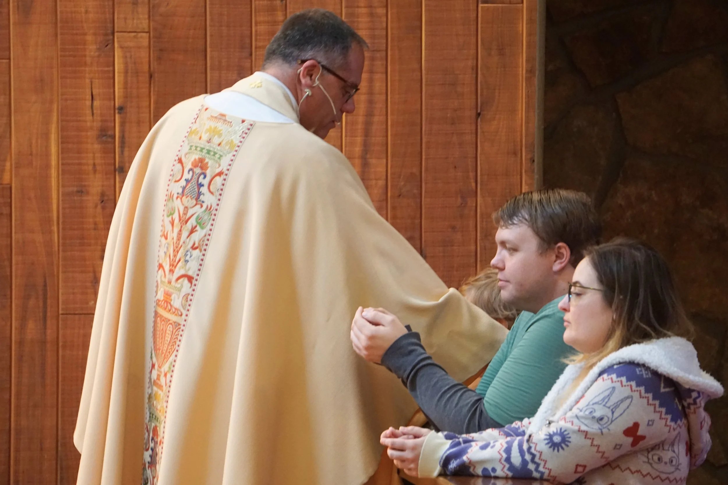 A priest wearing a cream-colored vestment with colorful embroidery is kneeling and placing his hand on the hand of a young woman with glasses, while other young women sit nearby inside a wooden chapel.