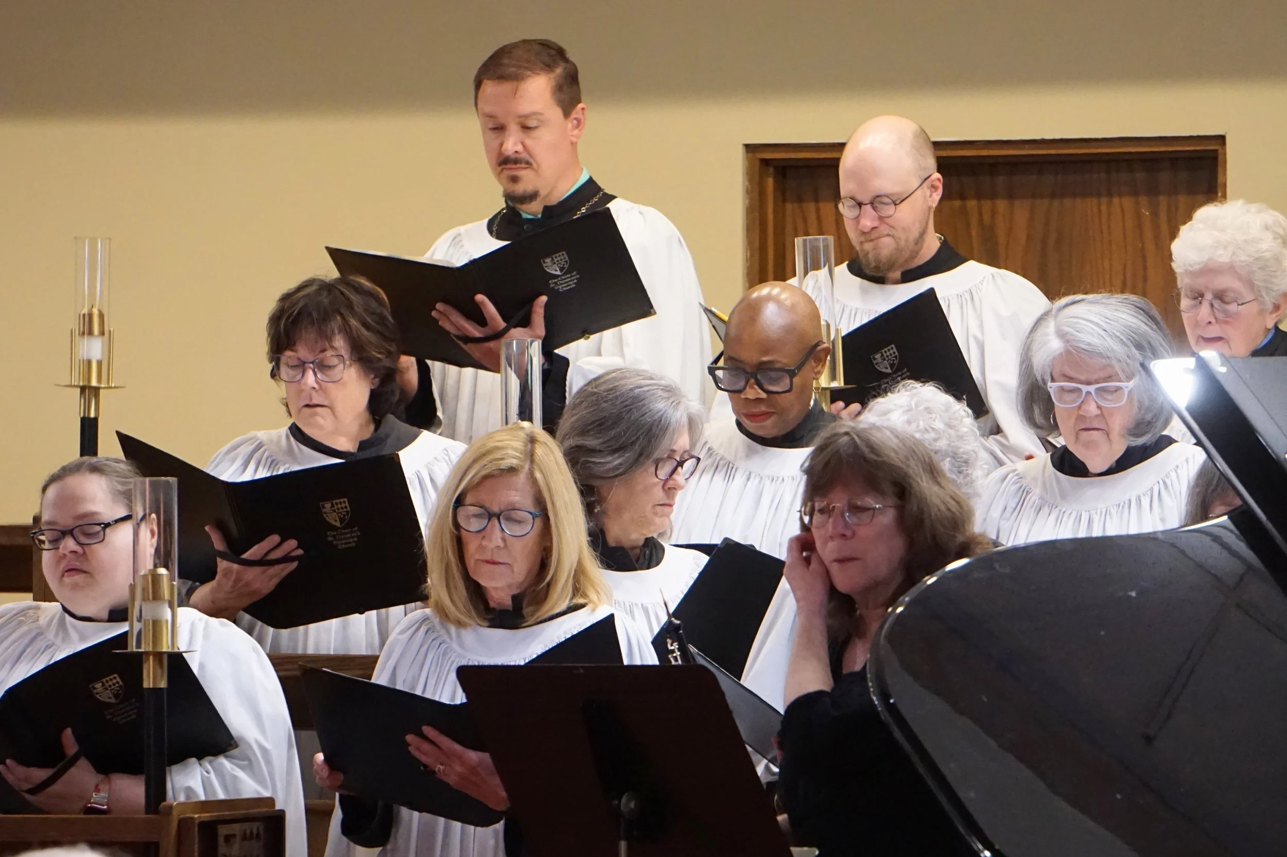 Choir members singing during a church service, with some reading sheet music, some holding music folders, and a few wearing glasses, in a church setting.
