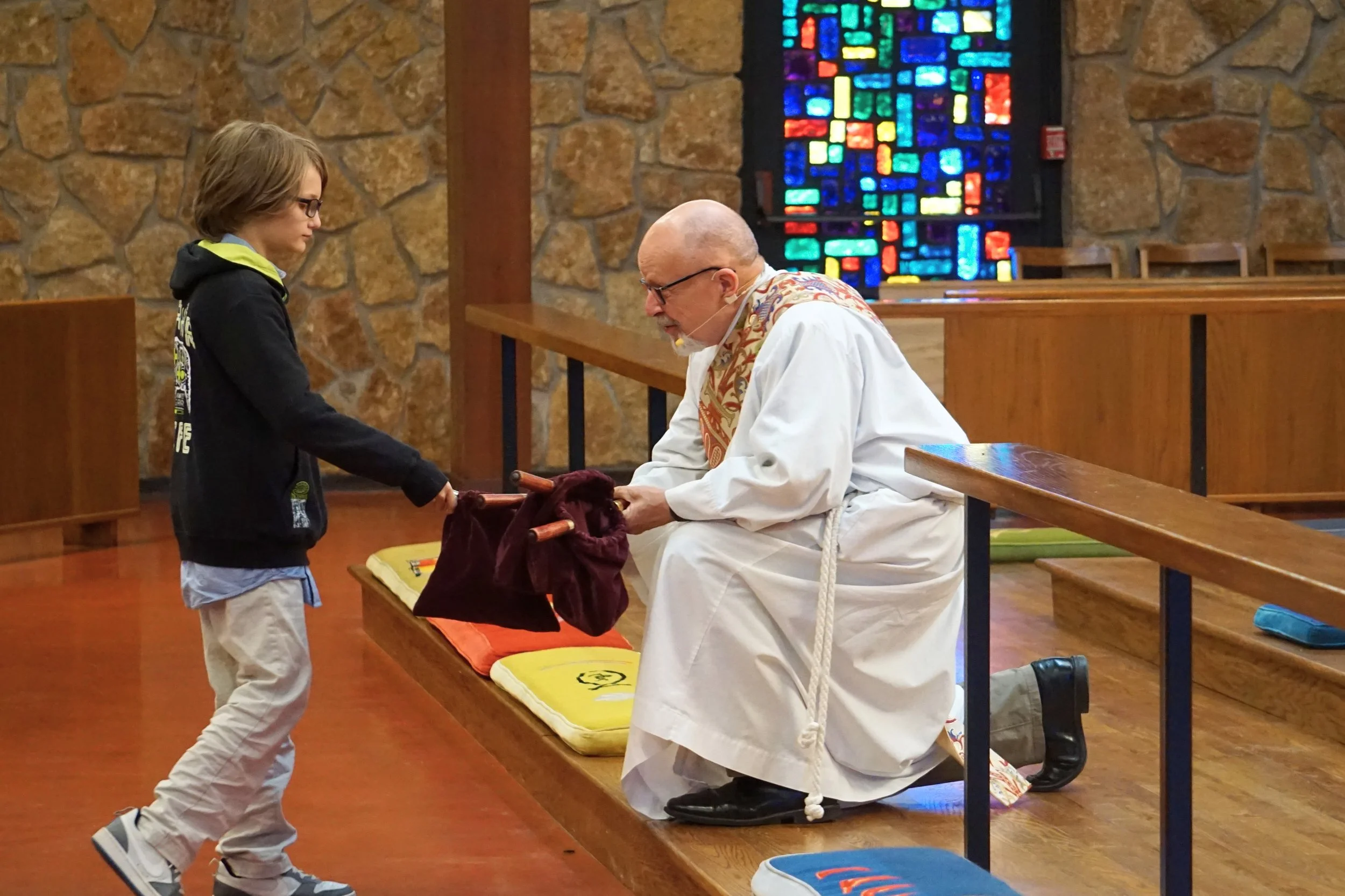 A young boy in a black hoodie and beige pants kneels before a priest in a white robe inside a church. They are engaged in a religious moment, with the boy holding a small object and the priest kneeling on a cushioned surface, with a stained-glass win