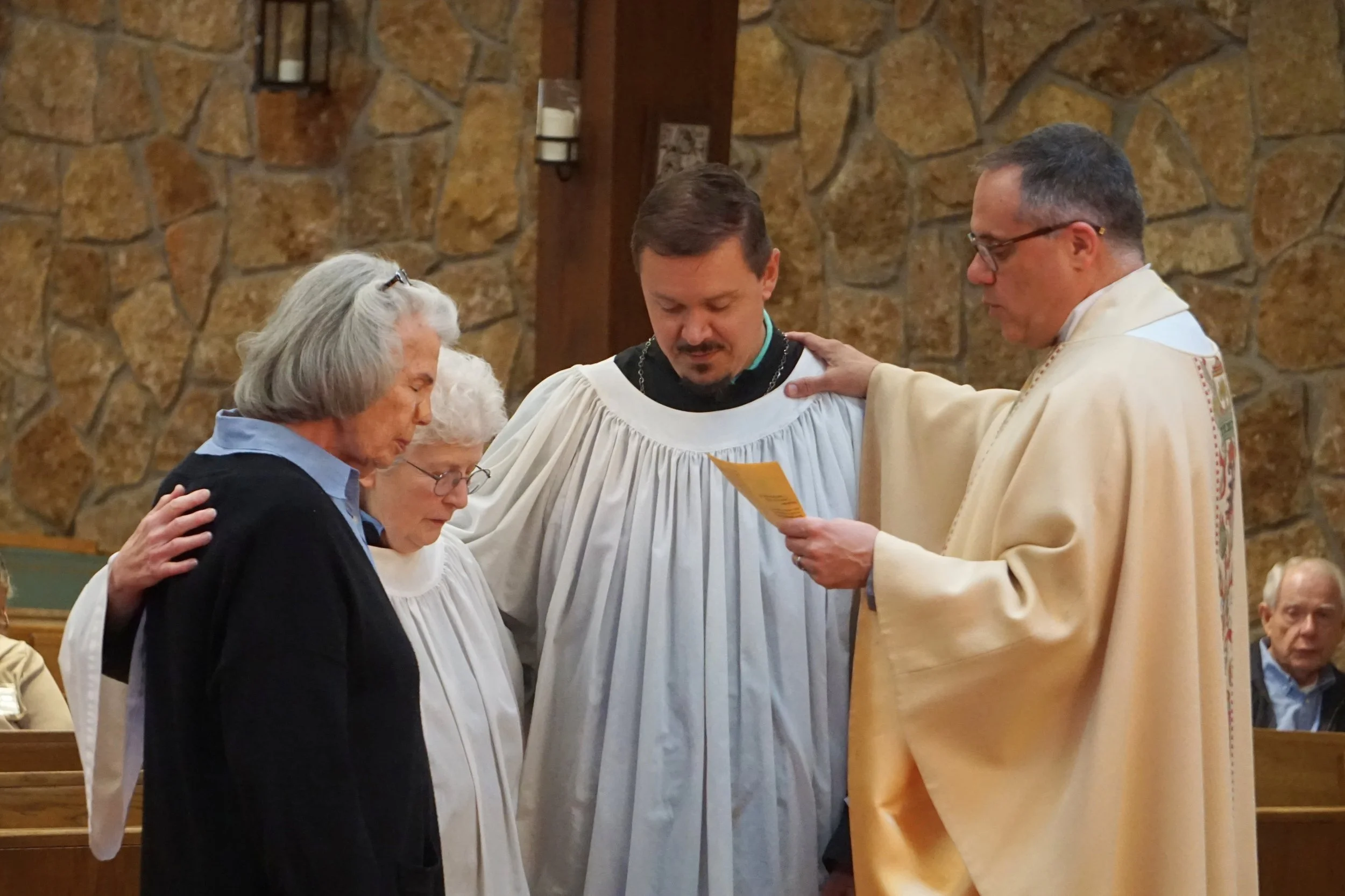 A priest in a white robe performing a religious ceremony with four women kneeling before him, one with her head bowed and hands clasped, in a church with a stone wall background.