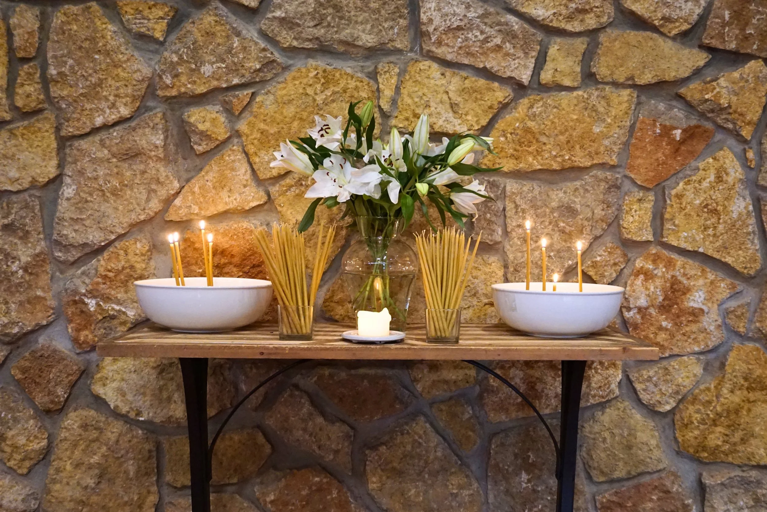 Flower arrangement with white lilies in a glass vase, surrounded by lit candles and incense sticks on a wooden shelf against a stone wall background.