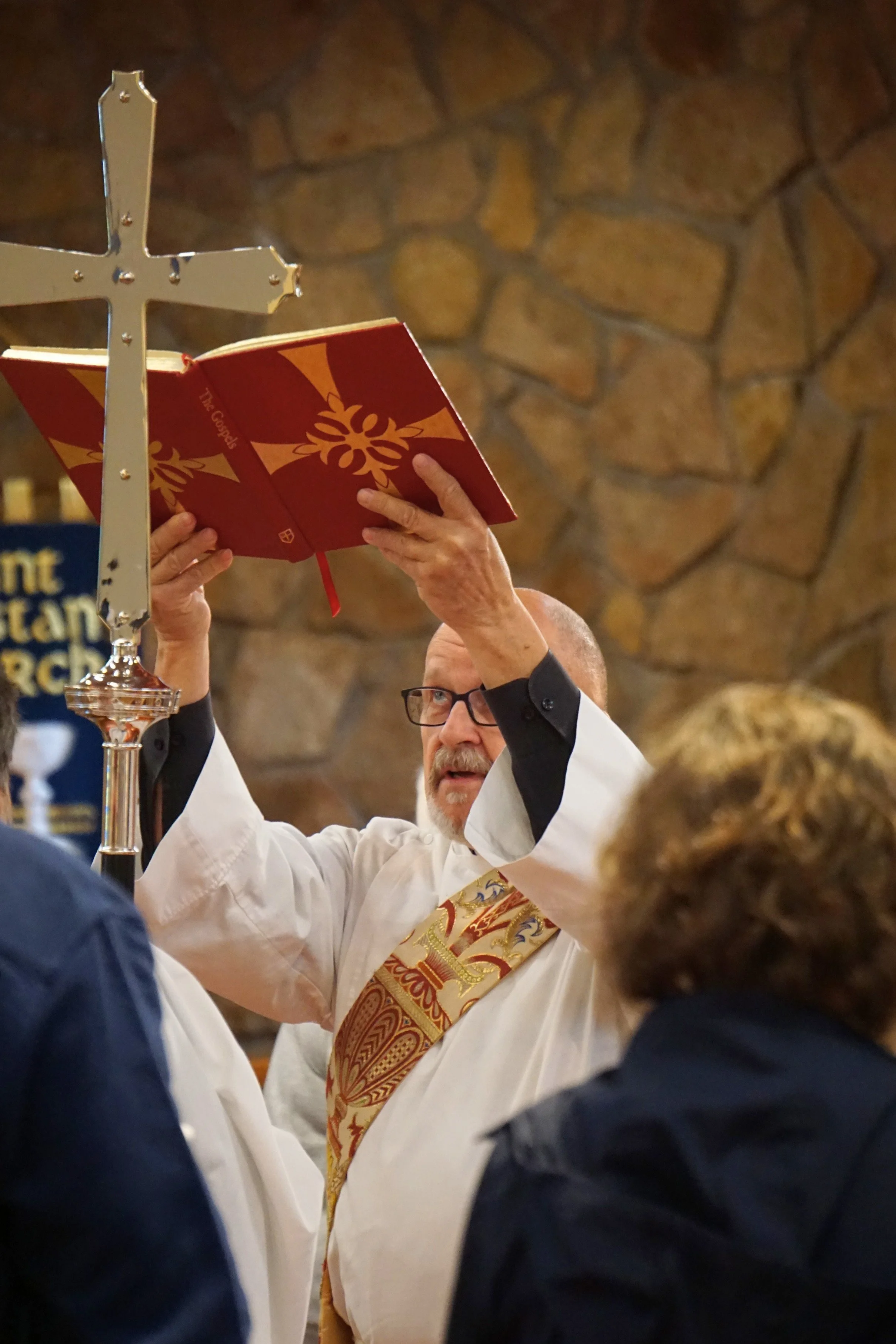 A priest with glasses is conducting a religious service, holding a red book titled 'The Gospel' in front of a stone wall background. Other people with dark and blonde hair are visible watching the service.