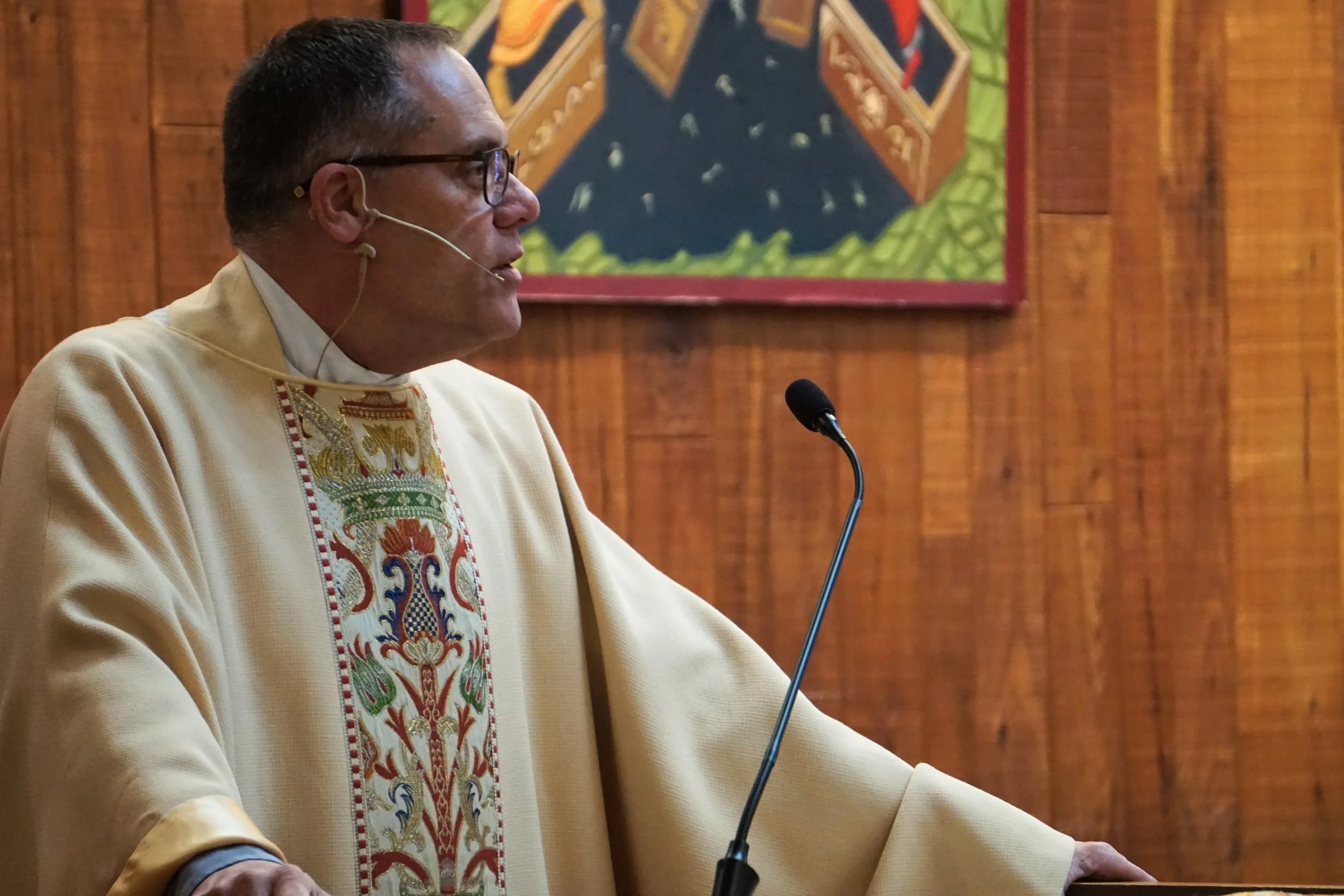 A man in traditional attire with glasses and a headset speaking at a podium with a microphone, in front of a wooden backdrop and colorful artwork on the wall.