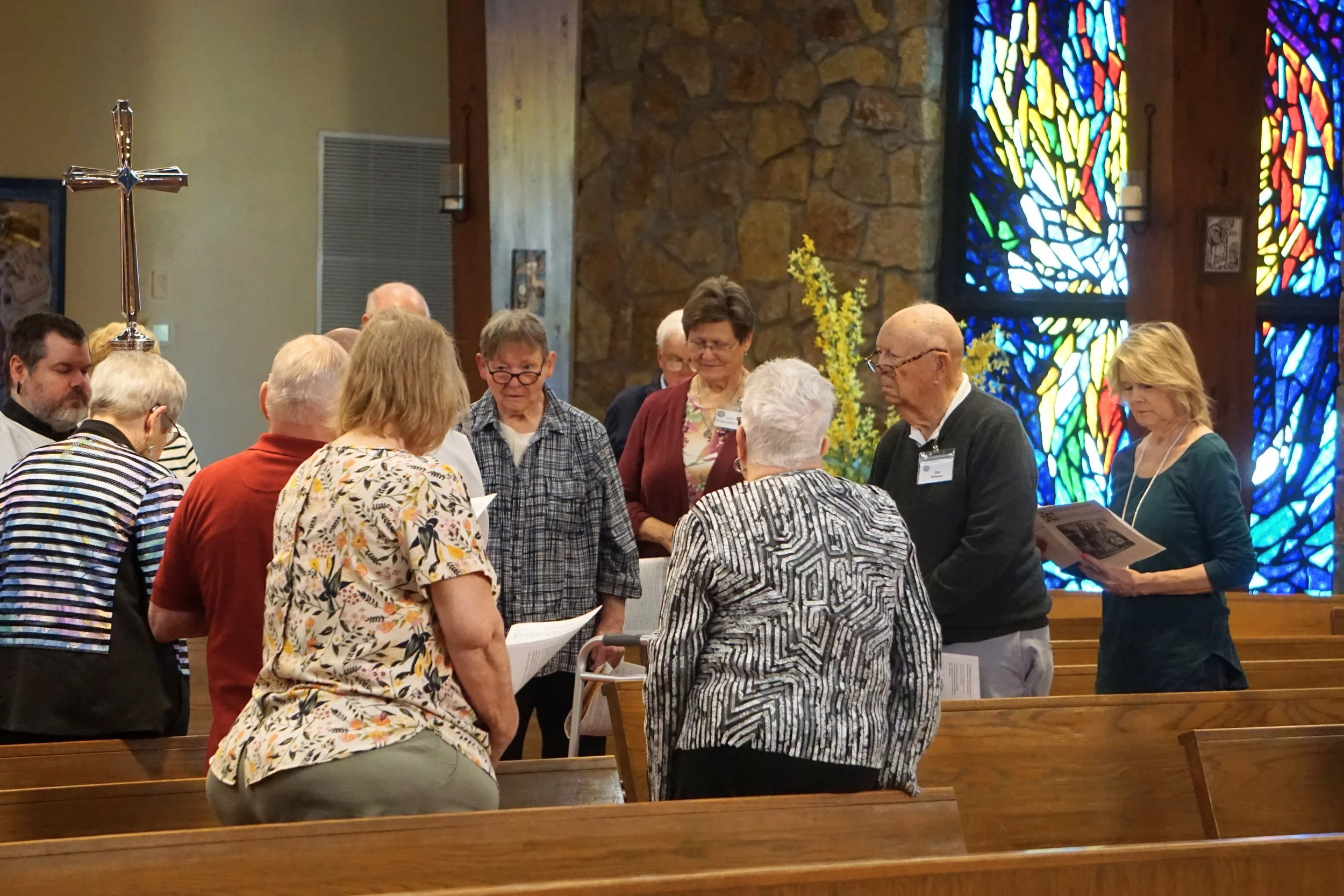A group of people gathered inside a church, praying or reading, with stained glass windows and wooden pews in the background.