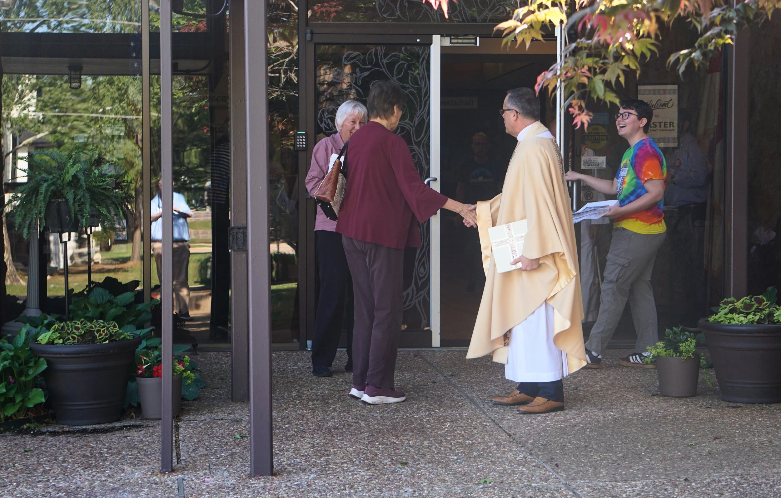 A group of people gathered outside the entrance of a building, including a person in a yellow graduation gown shaking hands with an older woman. A young person with glasses, wearing a tie-dye shirt, is smiling and holding papers, while another person