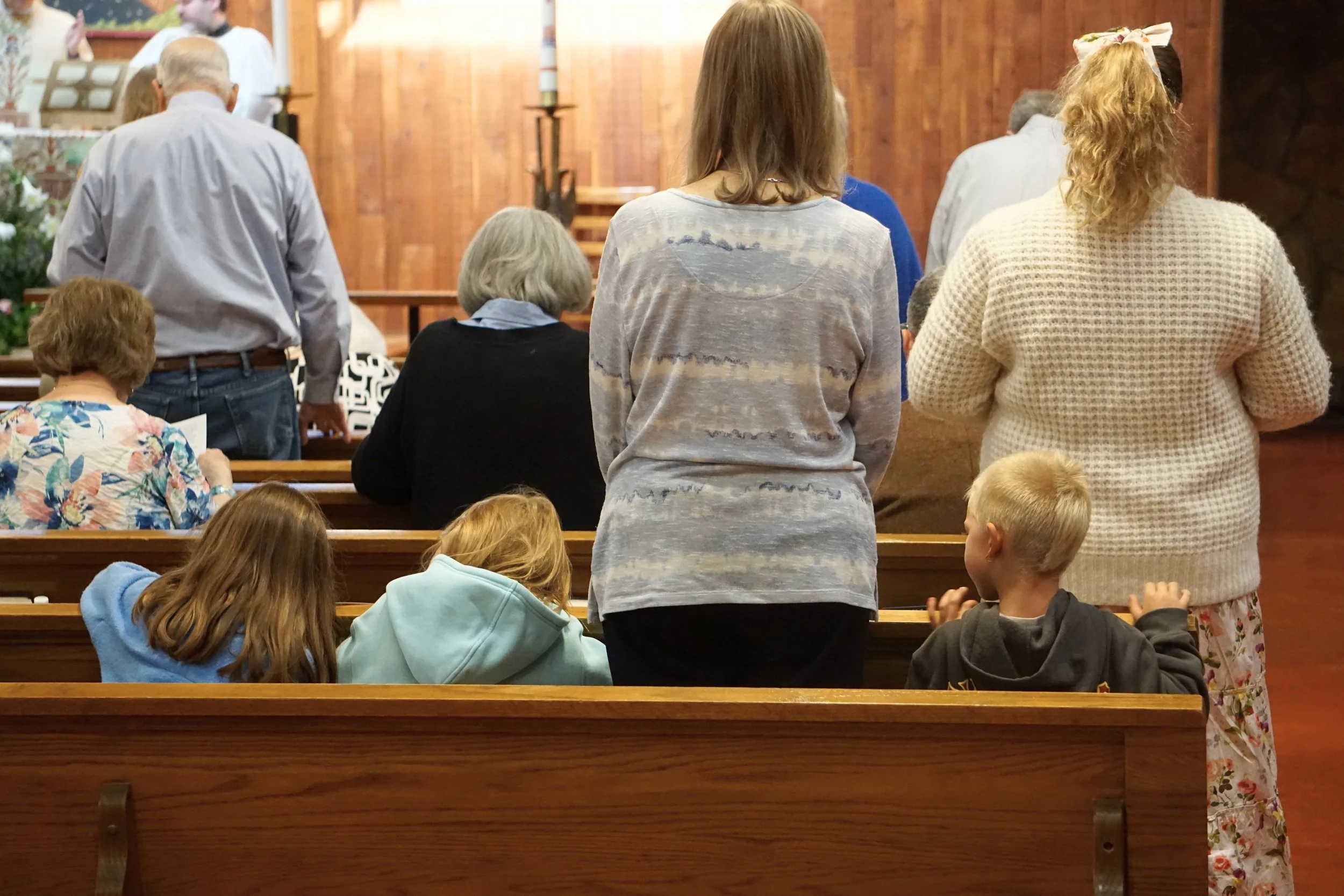 People praying in a church during a religious service, with some standing and some seated, wooden pews and a brick wall in the background.