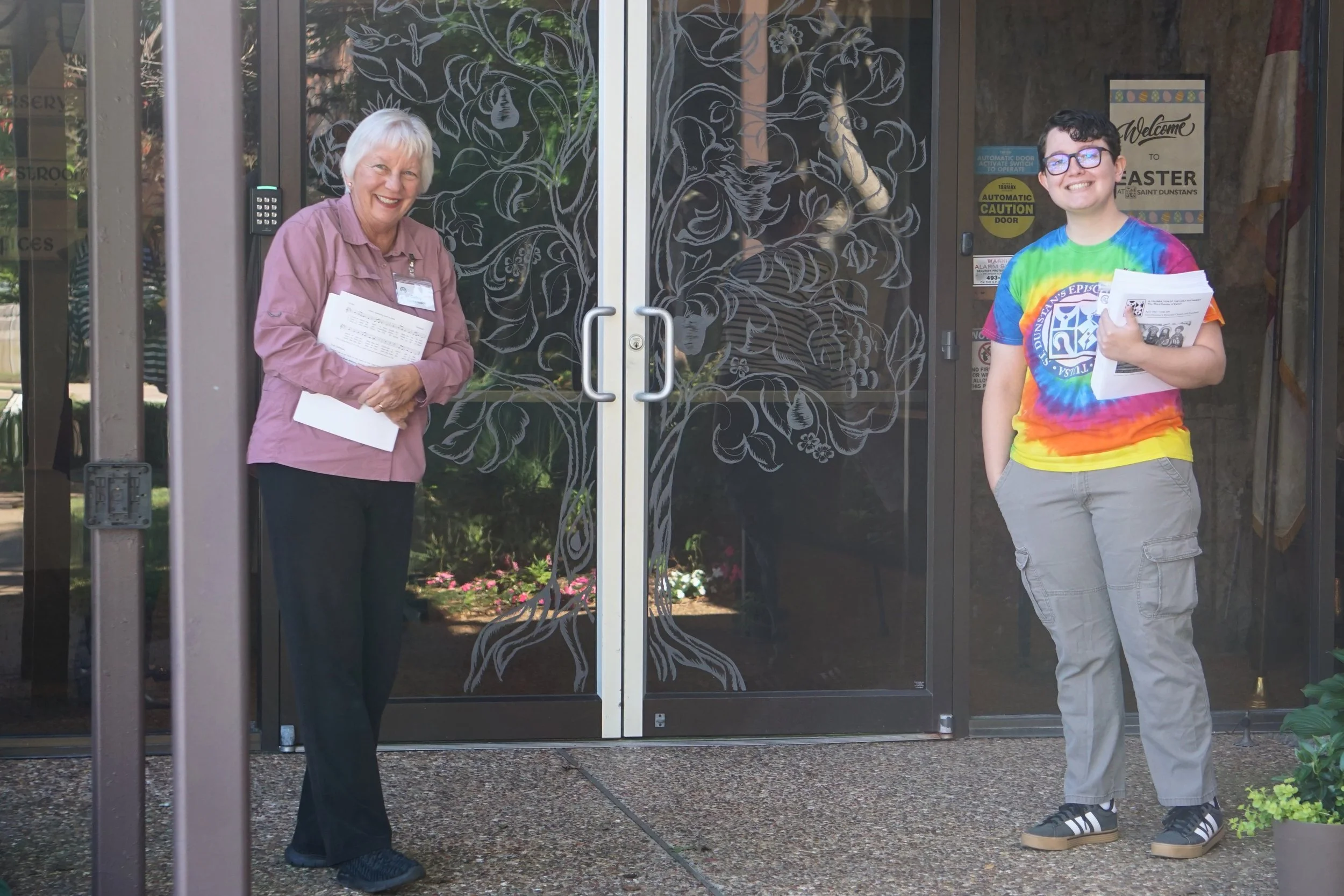Two women standing outside near glass doors decorated with floral patterns, holding papers, smiling, one wearing a pink shirt and the other in a tie-dye shirt.