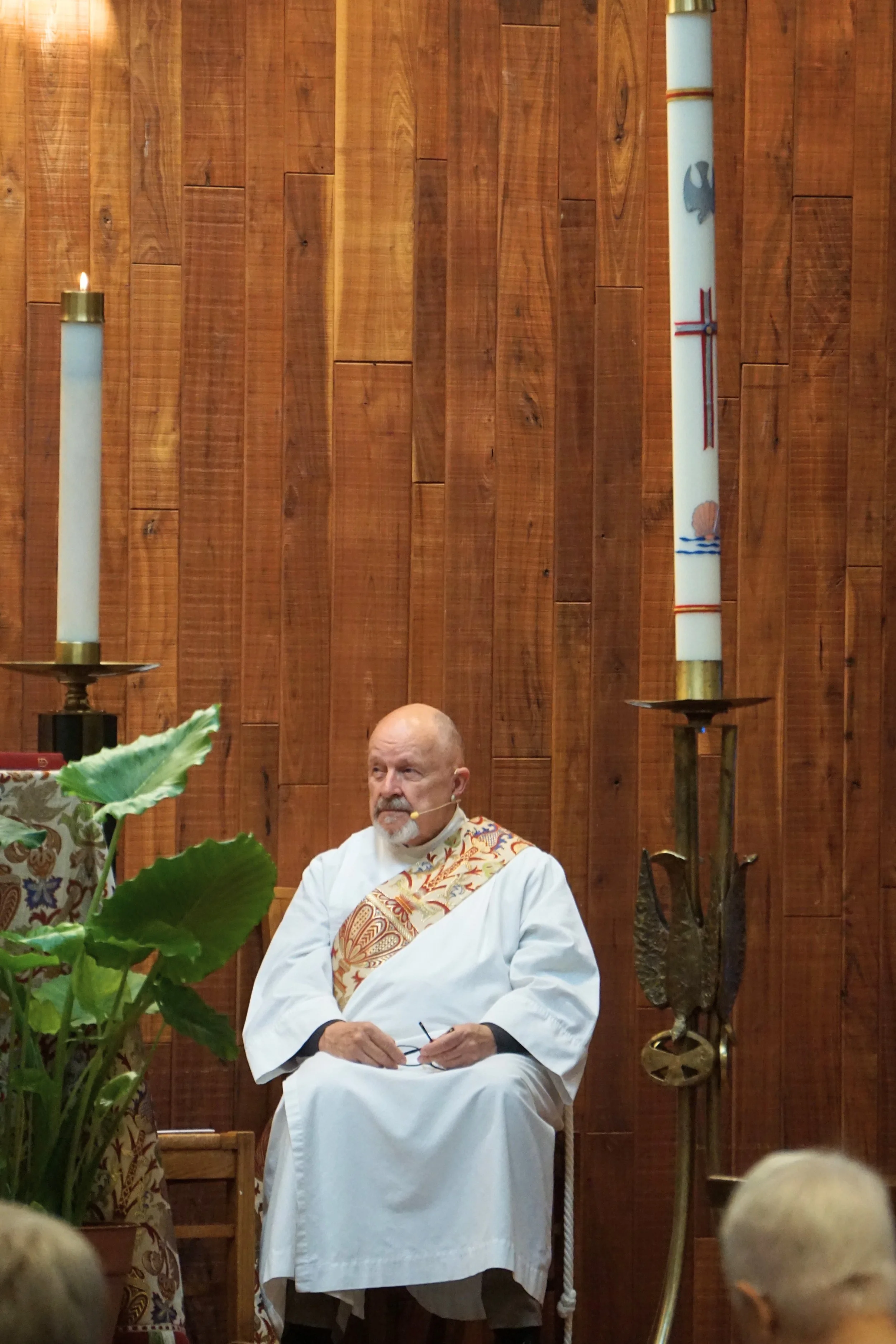 A man dressed in white religious robes sitting with arms crossed and holding glasses, in front of a wooden wall with large candles and religious symbols, possibly in a church or religious setting.