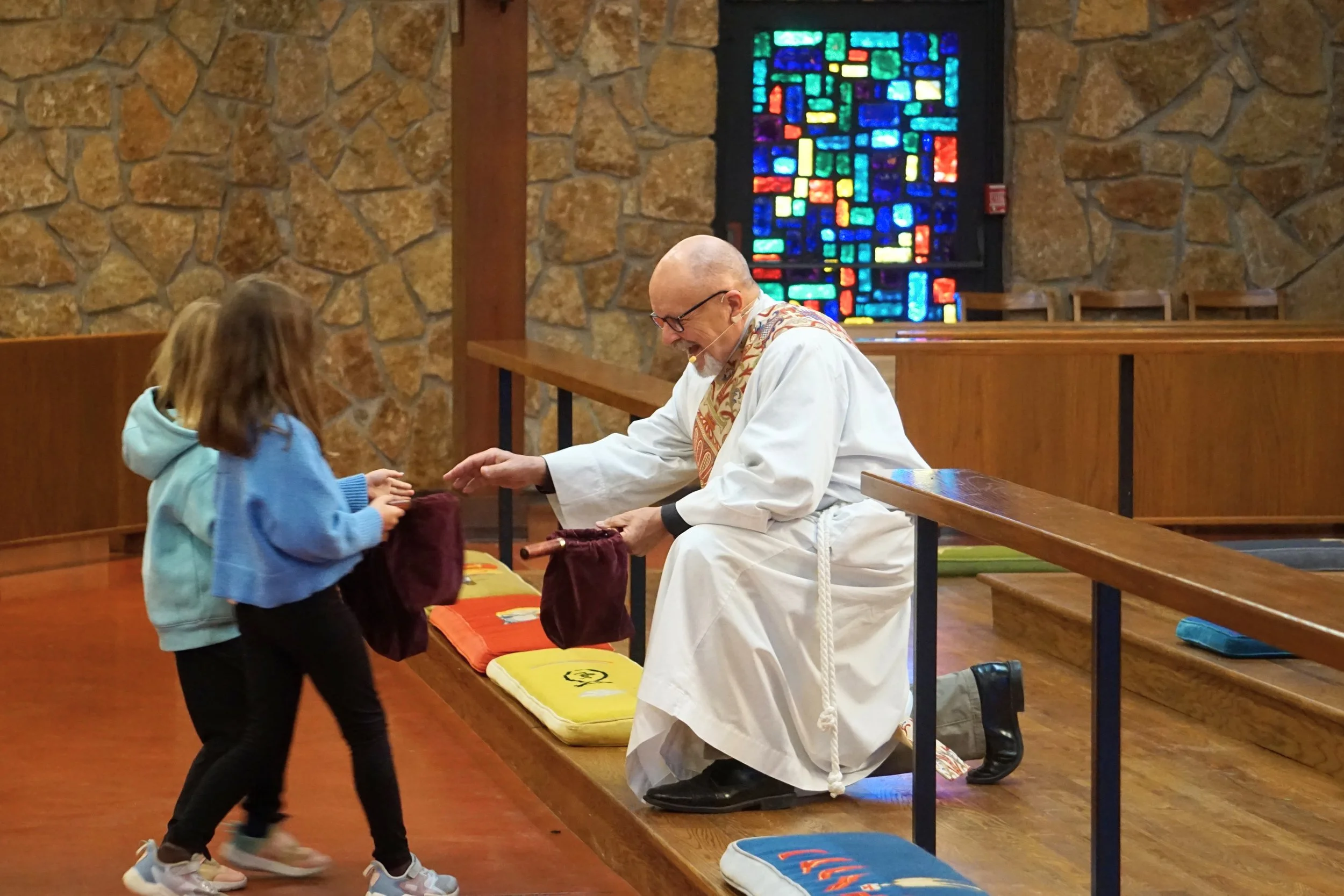 A priest dressed in white robes is sitting on a bench inside a church, smiling while receiving a gift from a young girl and her younger sibling. The girl is wearing a blue hoodie and the younger sibling is in a light blue jacket. Behind them is a col