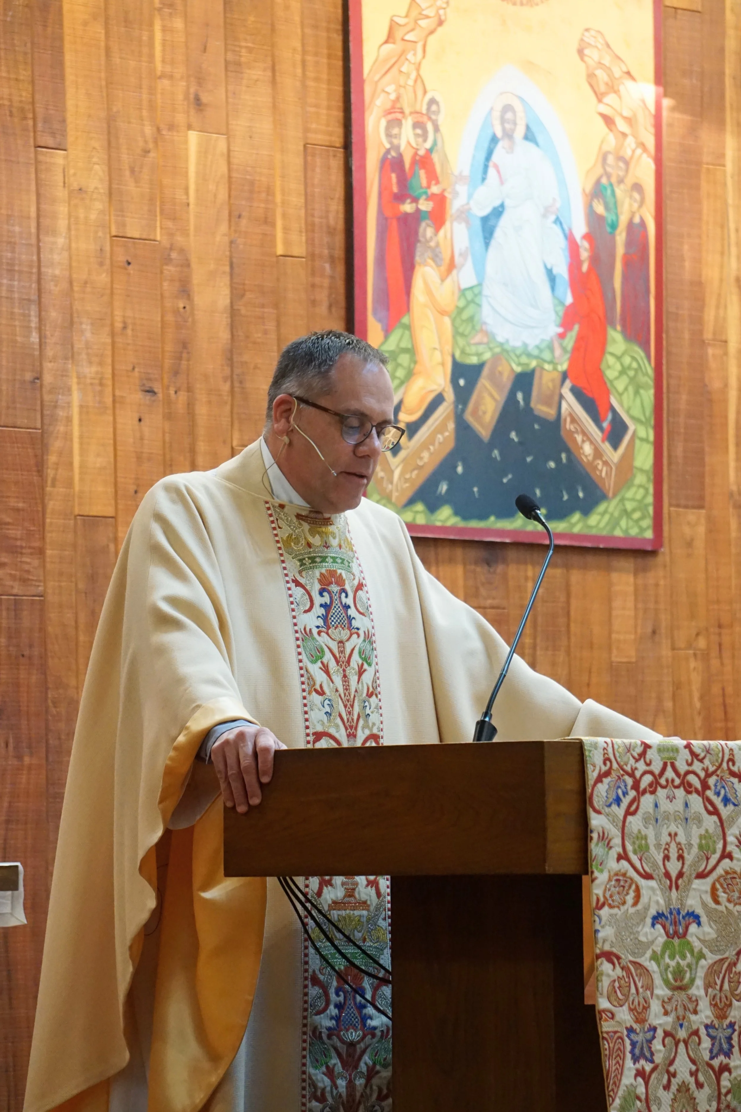 A priest in cream vestments standing at a wooden lectern, reading or speaking, with a mural of religious figures in the background.