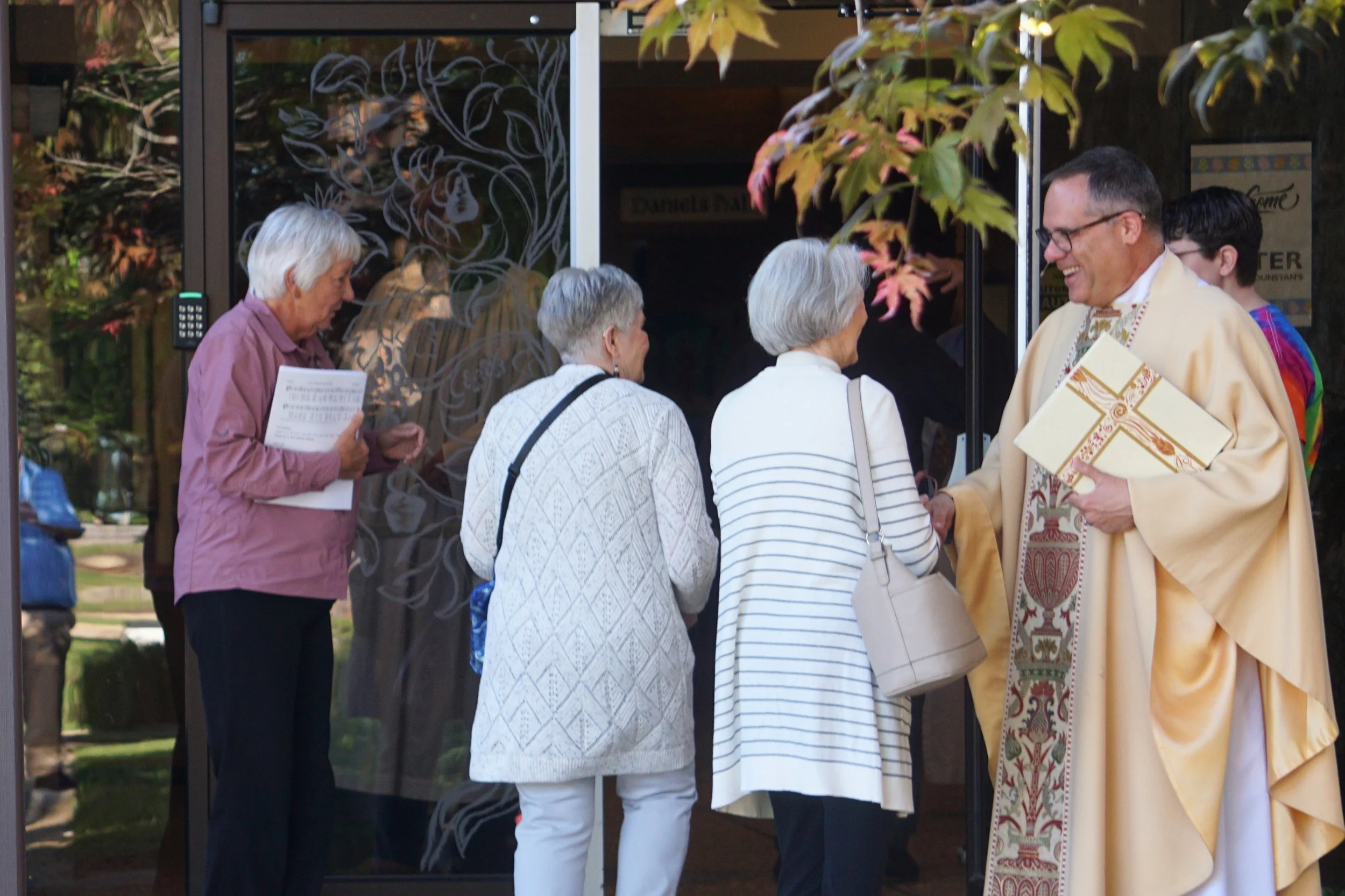 A group of five elderly people gathered outside a building, engaging in conversation, with a priest in ceremonial robes holding a book, during fall as indicated by the colorful autumn leaves and the warm clothing.