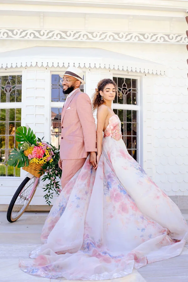 A couple dressed in elegant, pastel-colored wedding attire standing back-to-back in front of a white house with decorative trim. The bride's flowing gown features watercolor patterns, and the groom wears a light pink suit and a fedora hat. A yellow b