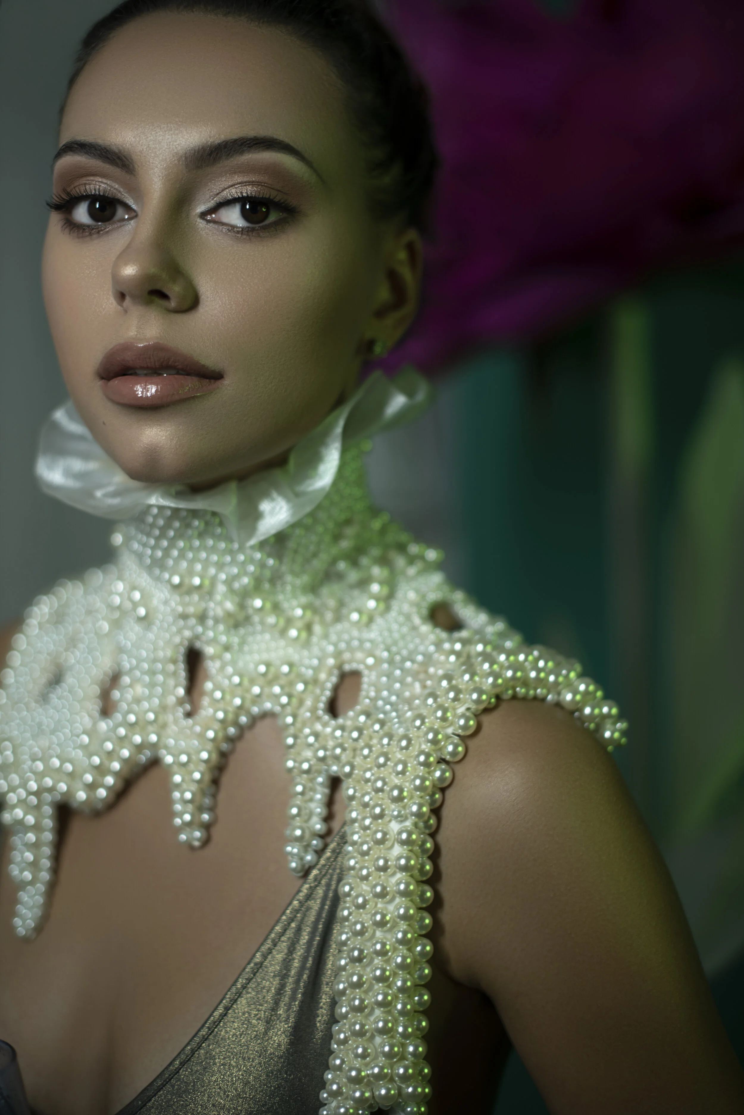 A woman with flawless makeup and shimmering lipstick, wearing a pearl-embellished top with a high ruffled collar, and a metallic dress, against a blurred dark background with purple flowers.