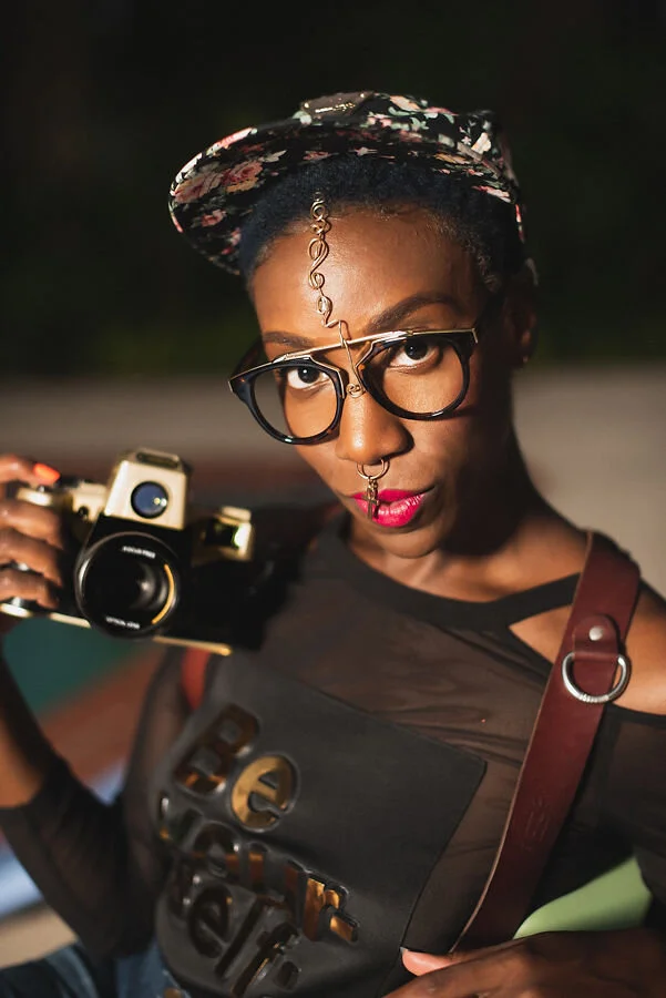 A woman wearing glasses and a floral hat holding a camera, with a confident expression at night.