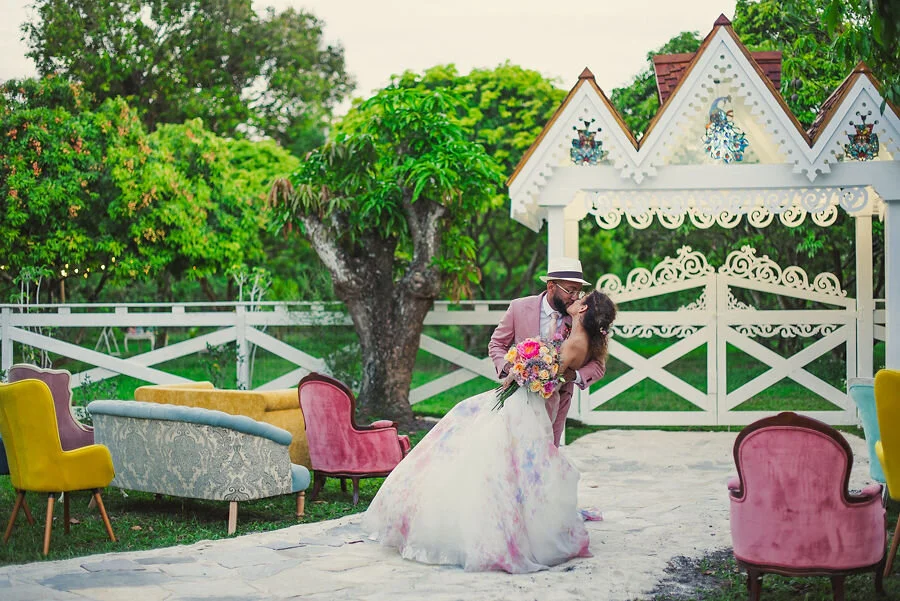A newlywed couple shares a kiss outdoors amidst colorful vintage chairs, with a decorative white gazebo and green trees in the background.