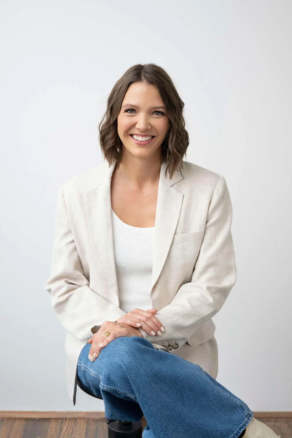 A smiling woman with shoulder-length brown hair wearing a light beige blazer, white top, and blue jeans, sitting with her hands crossed on her lap against a plain light background.