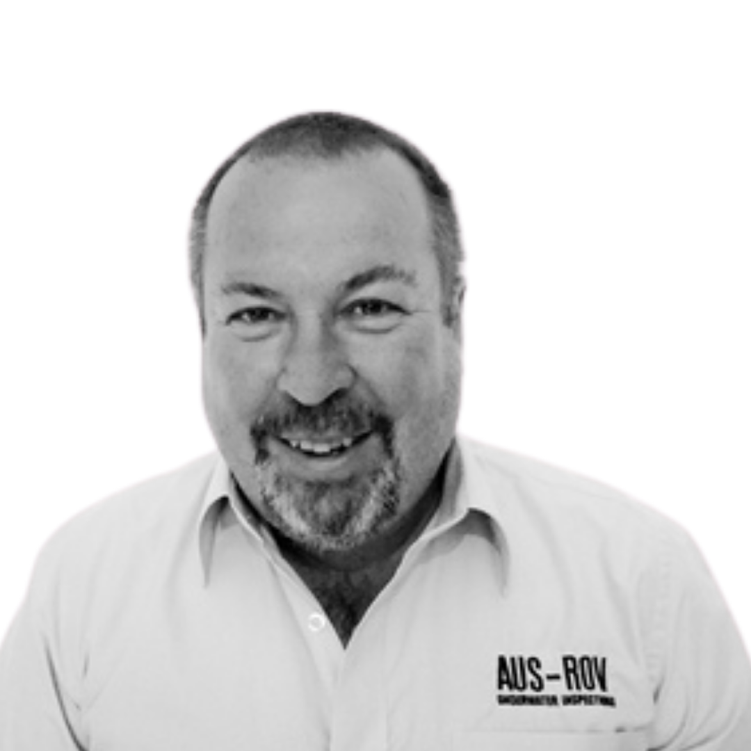 A smiling man with short hair and a beard, wearing a collared shirt with the logo 'AUS-ROY' on it.