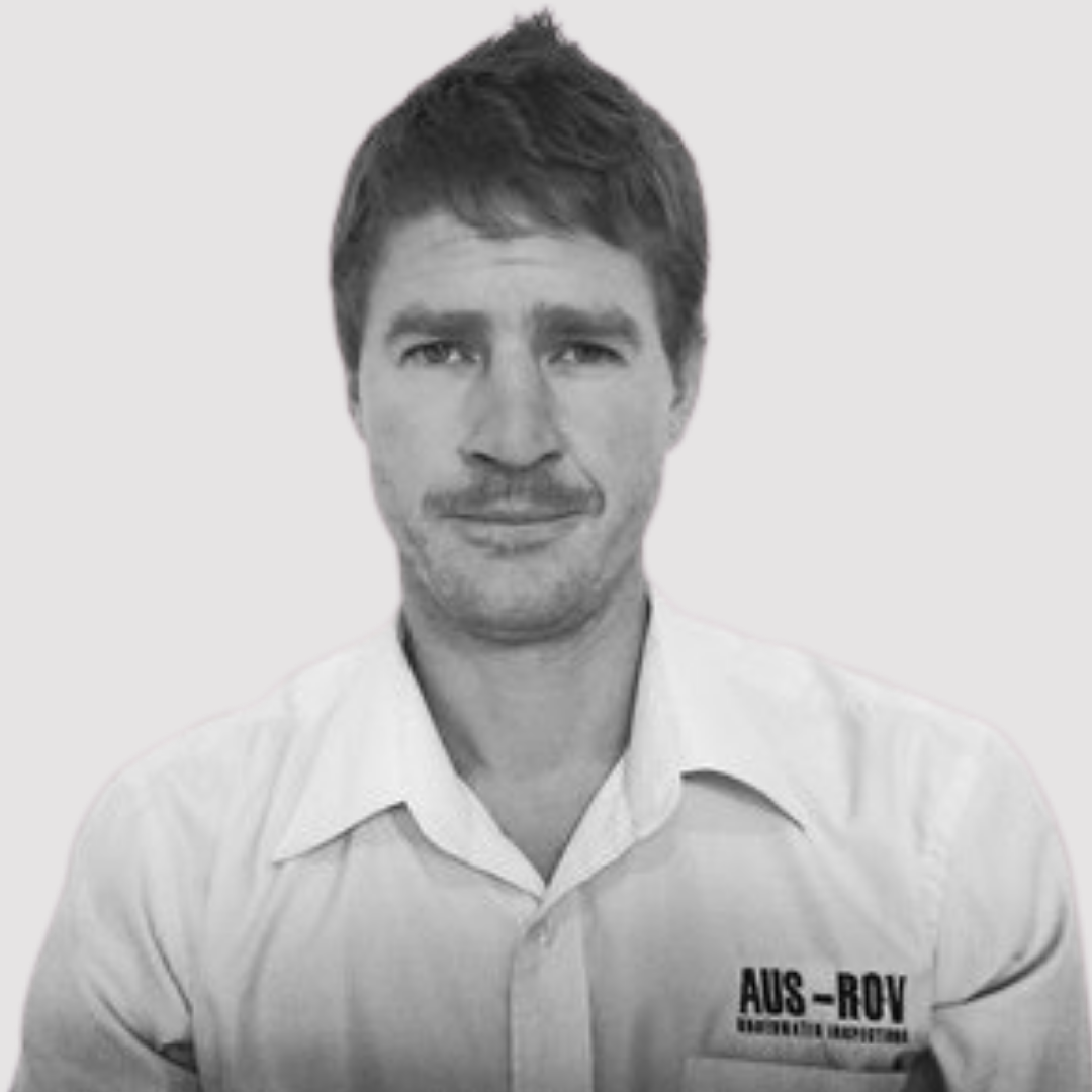 Black and white photo of a man with short, slightly messy hair, wearing a collared shirt with a logo or text on the left chest.