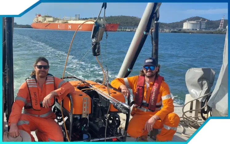 Two men wearing orange survival suits and sunglasses on a boat, with a large cargo ship and industrial buildings in the background.