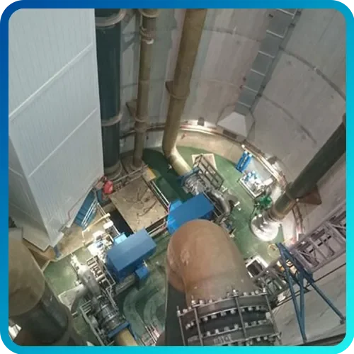 View looking down at a large industrial machine inside a cylindrical concrete structure, with pipes and equipment surrounding it.