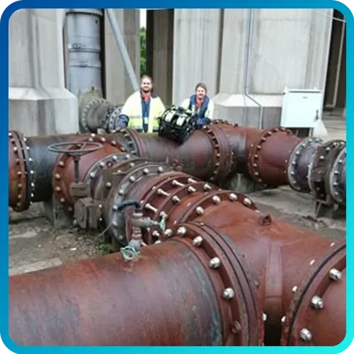 Two workers in safety vests standing behind large rusted industrial pipes outdoors at a facility.