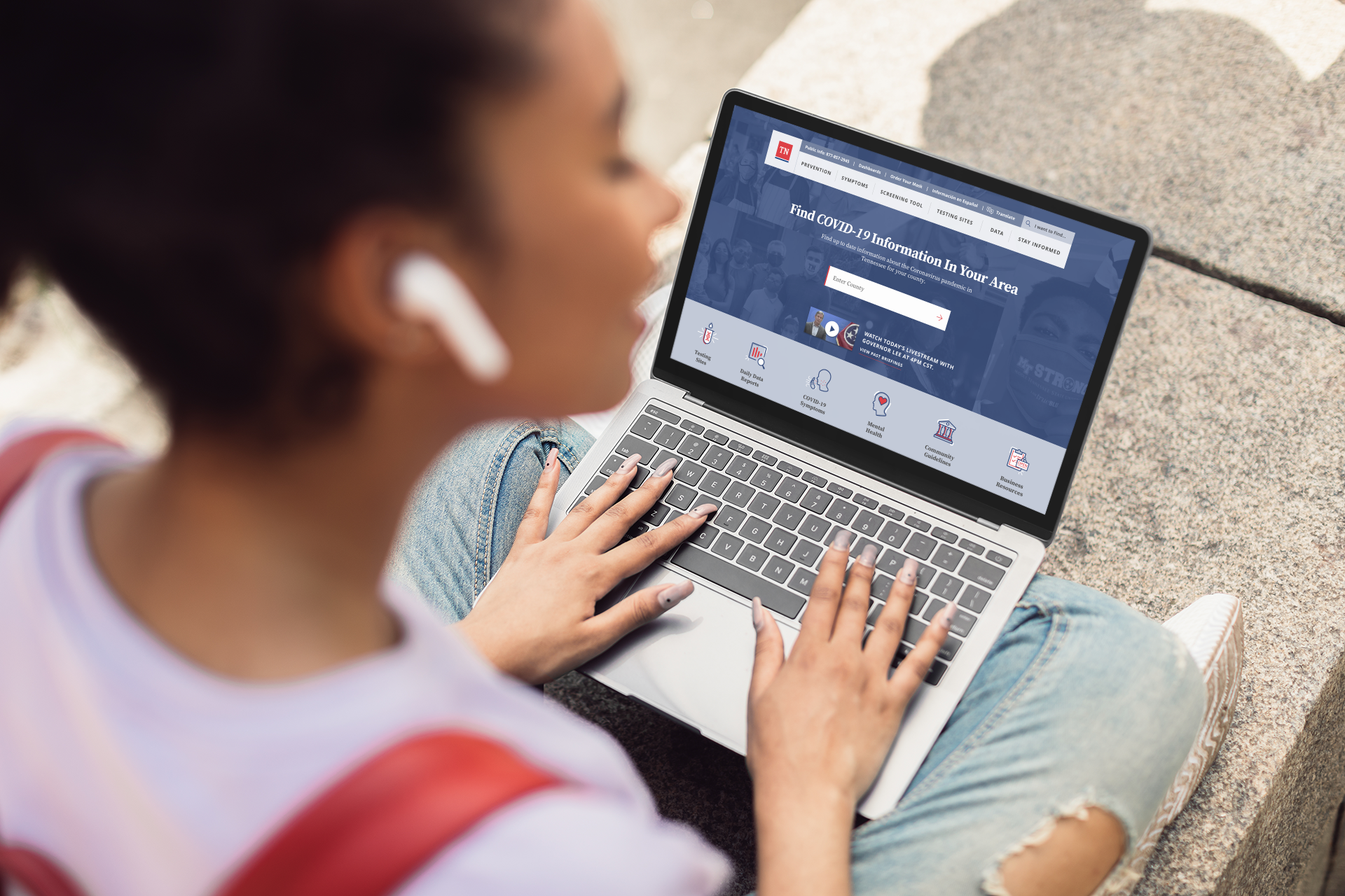  Woman wearing headphones and a backpack using the COVID-19 Information Hub website on her laptop while she sits cross legged.  