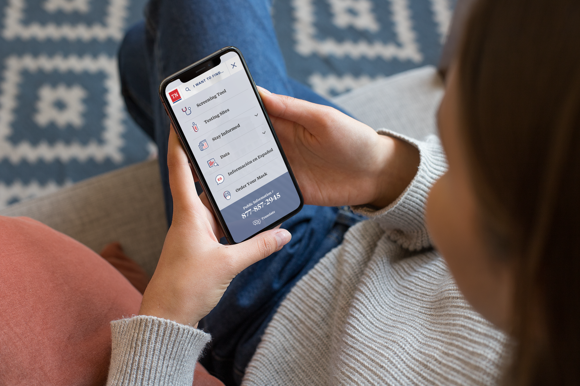  Woman wearing a sweater using the COVID-19 Information Hub on her iPhone while sitting on her couch.  