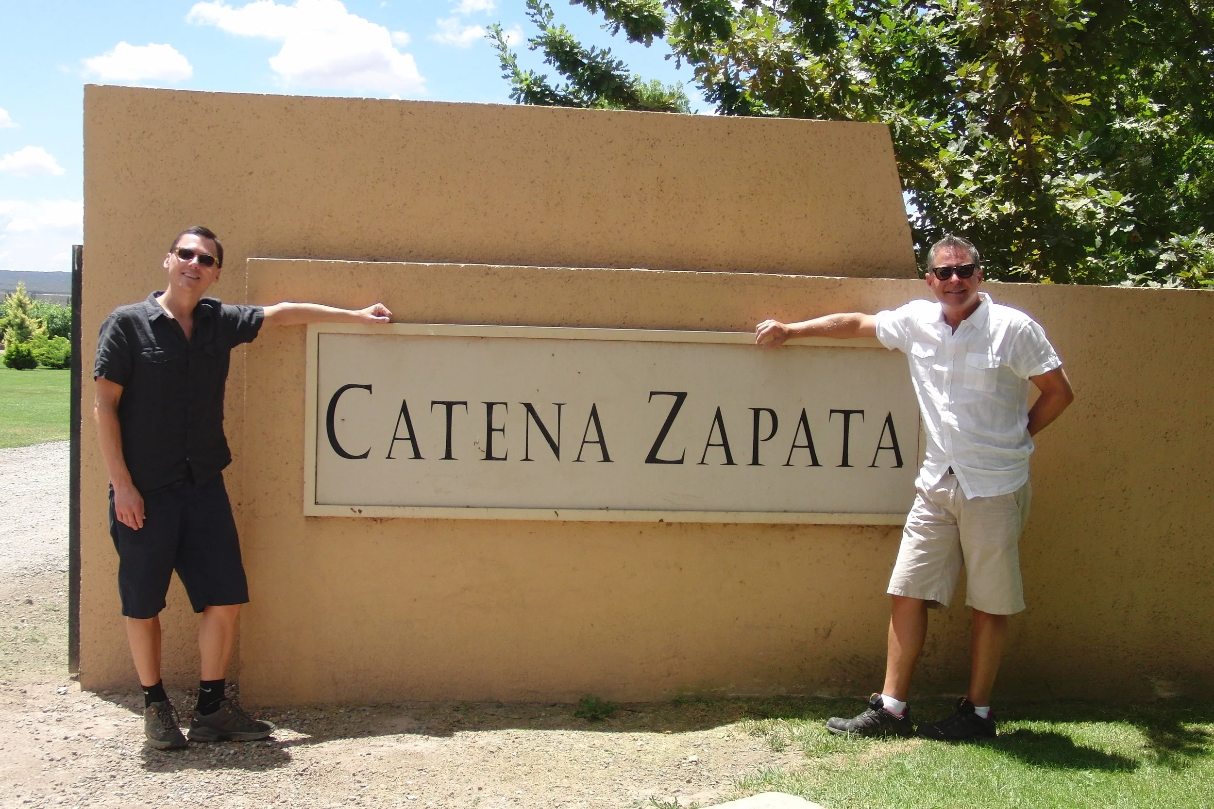 Two men wearing sunglasses stand next to a beige sign that reads 'Catenazapata' in black letters, outdoors with green trees and blue sky with clouds in the background.