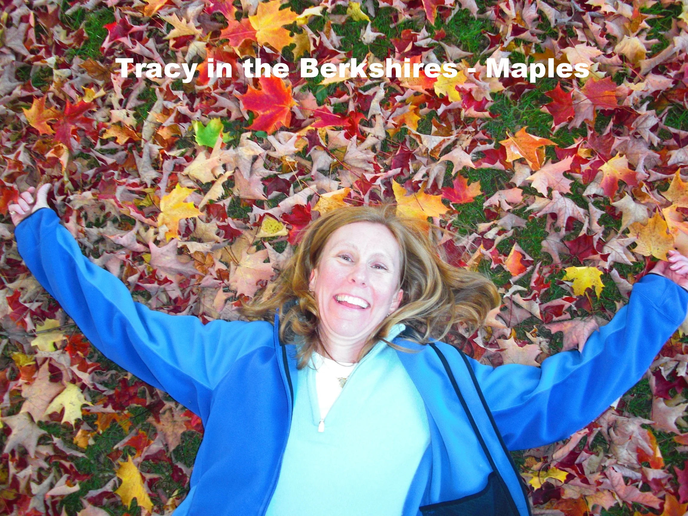 A woman with long red hair lying on the ground among colorful fallen autumn leaves, smiling and wearing a blue jacket.