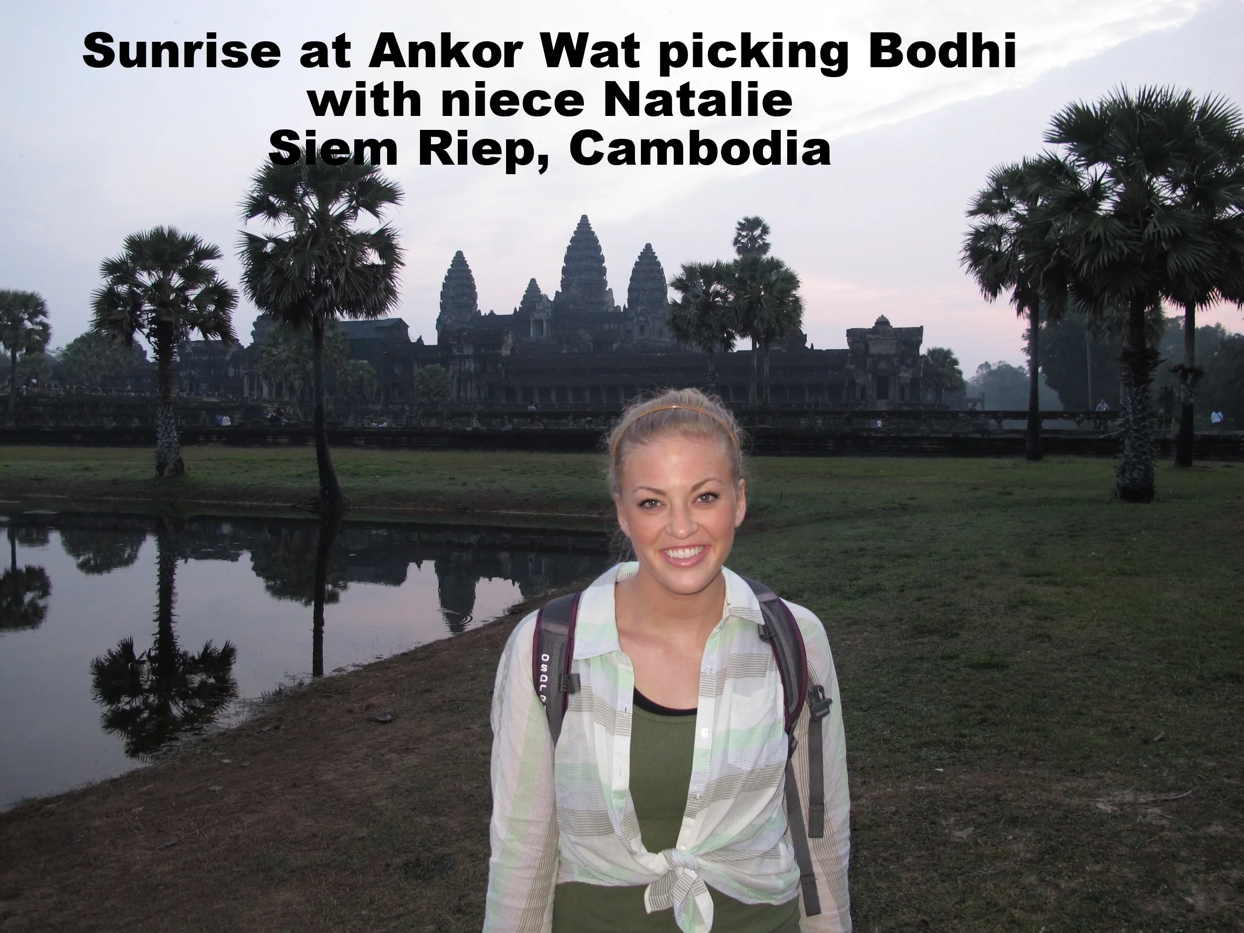 A woman with blonde hair smiling, standing in front of a pond with the ancient Angkor Wat temple in Siem Reap, Cambodia, during sunrise, with trees and a cloudy sky.